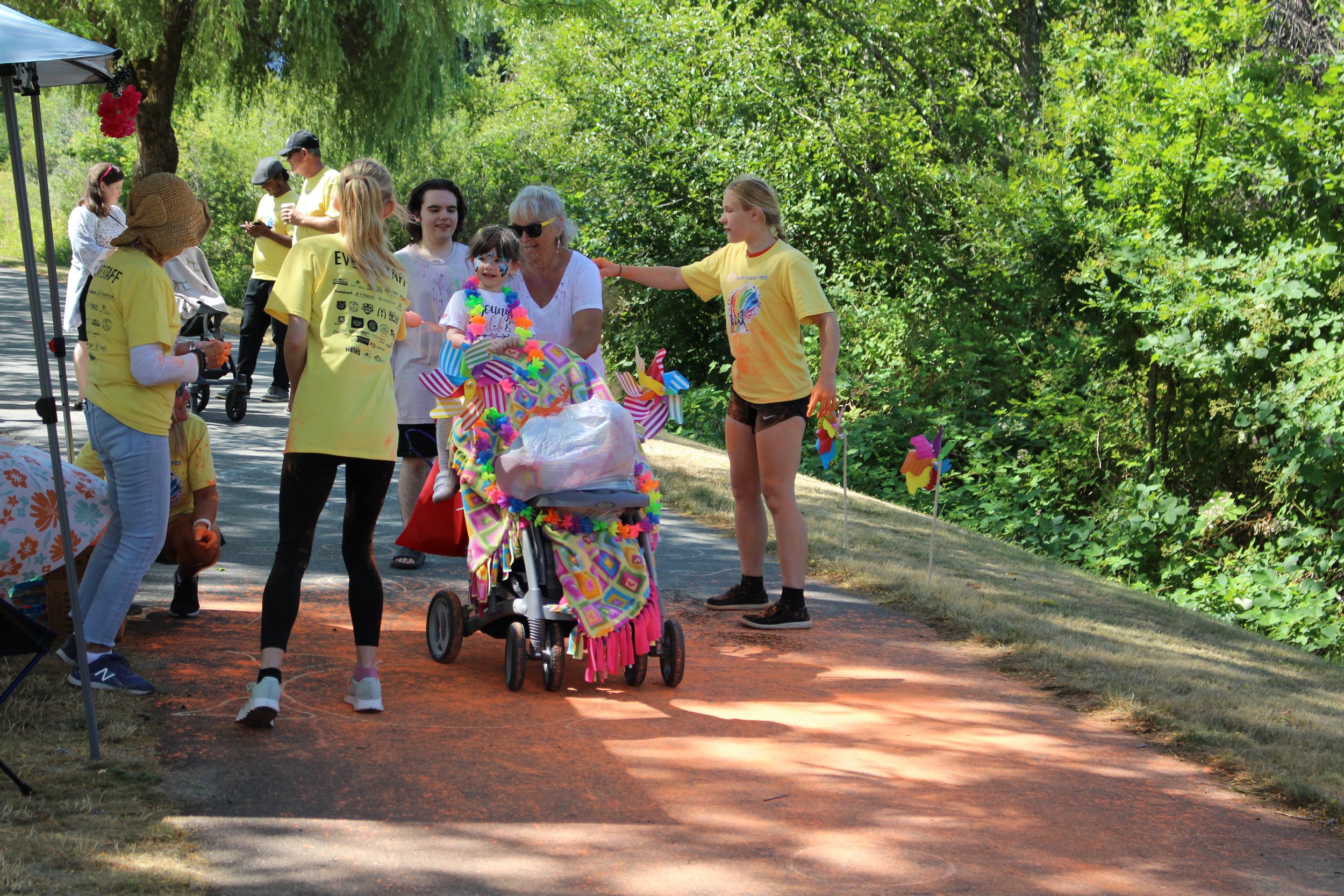 Group of people gathered around a colorful stroller decorated with presents and a clown-themed blanket, outdoors on a sunny day with green trees in the background.
