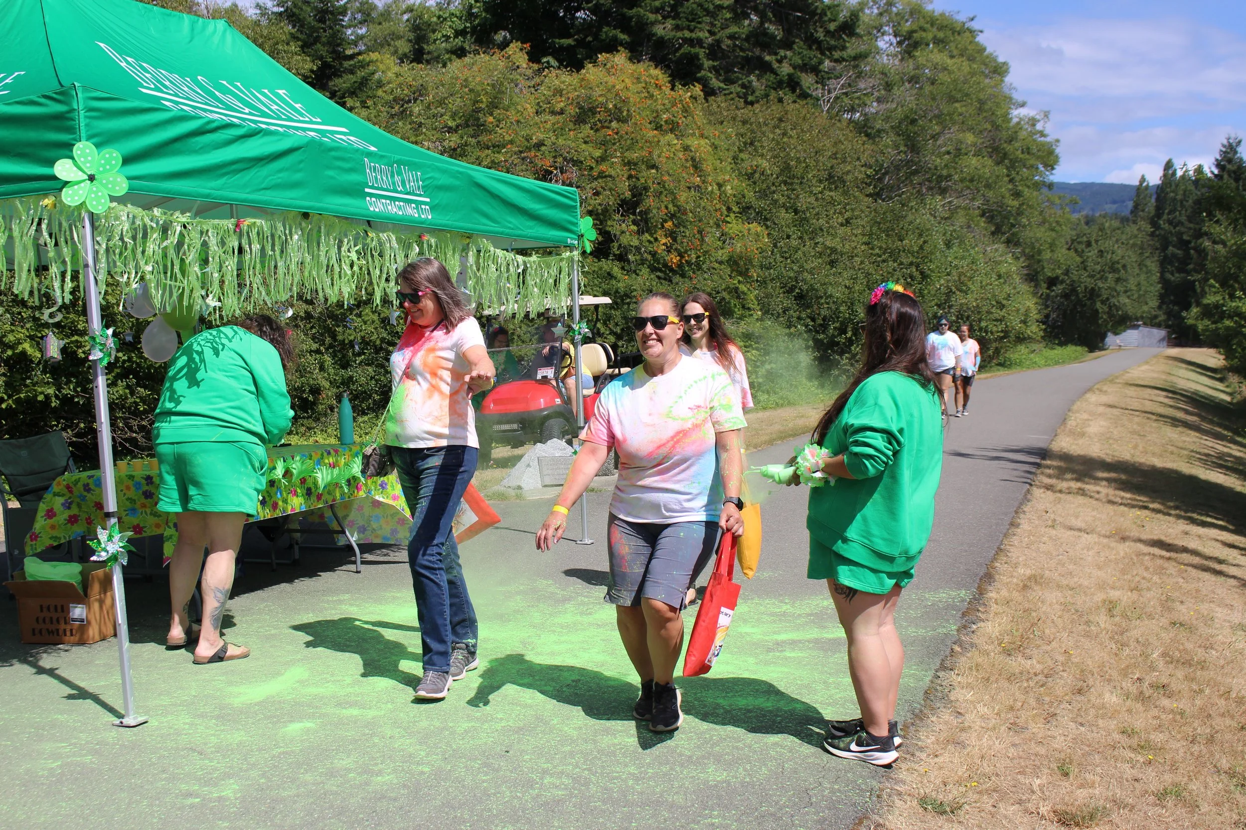 People participating in a color powder event near a green tent with a grassy outdoor setting and trees in the background.