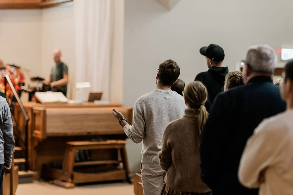 People standing and sitting in a church or gathering space, listening to a speaker at a pulpit with musical instruments in the background.