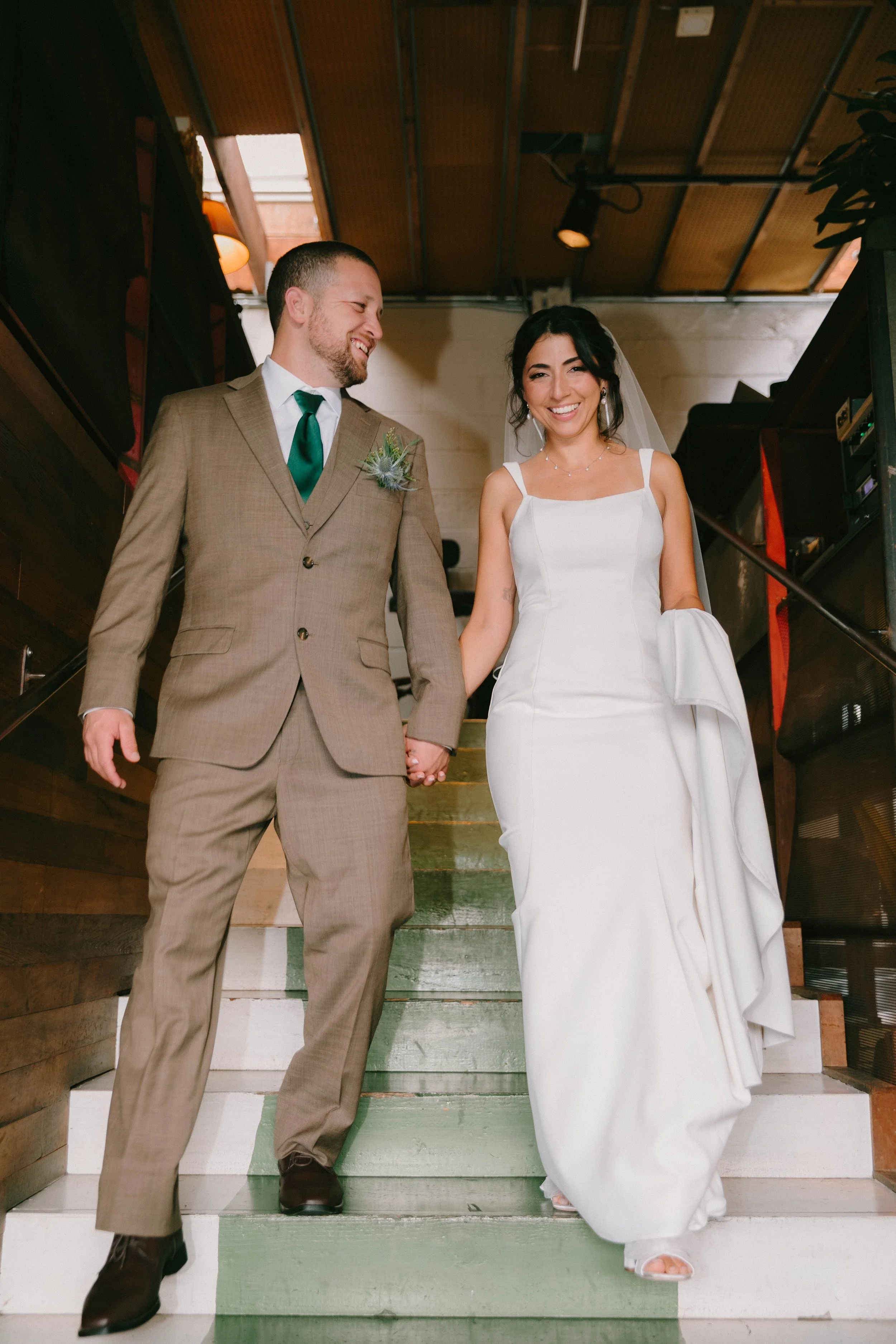 A newlywed couple holding hands and smiling down a set of painted green and white stairs inside a building.