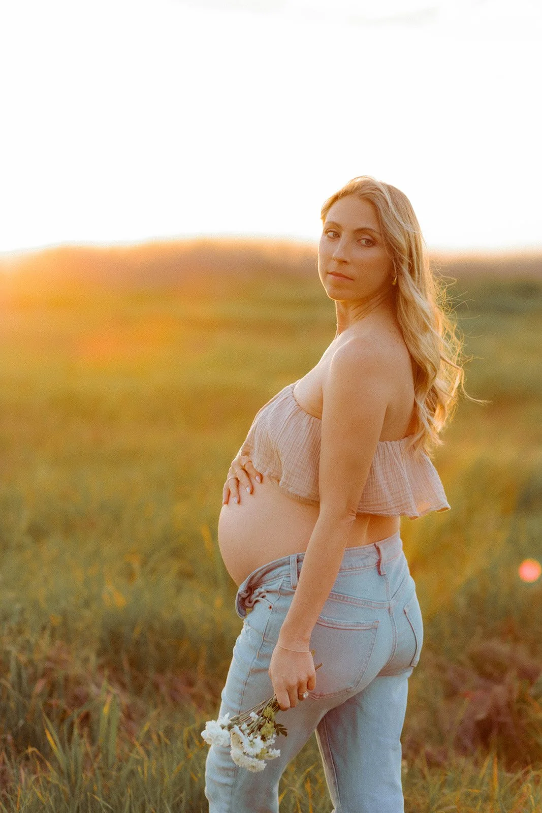 Pregnant mother looking back at camera holding belly at sunset in green, golden field in Costa Mesa — Best maternity photographers in Orange County — Tesla Rae Photo