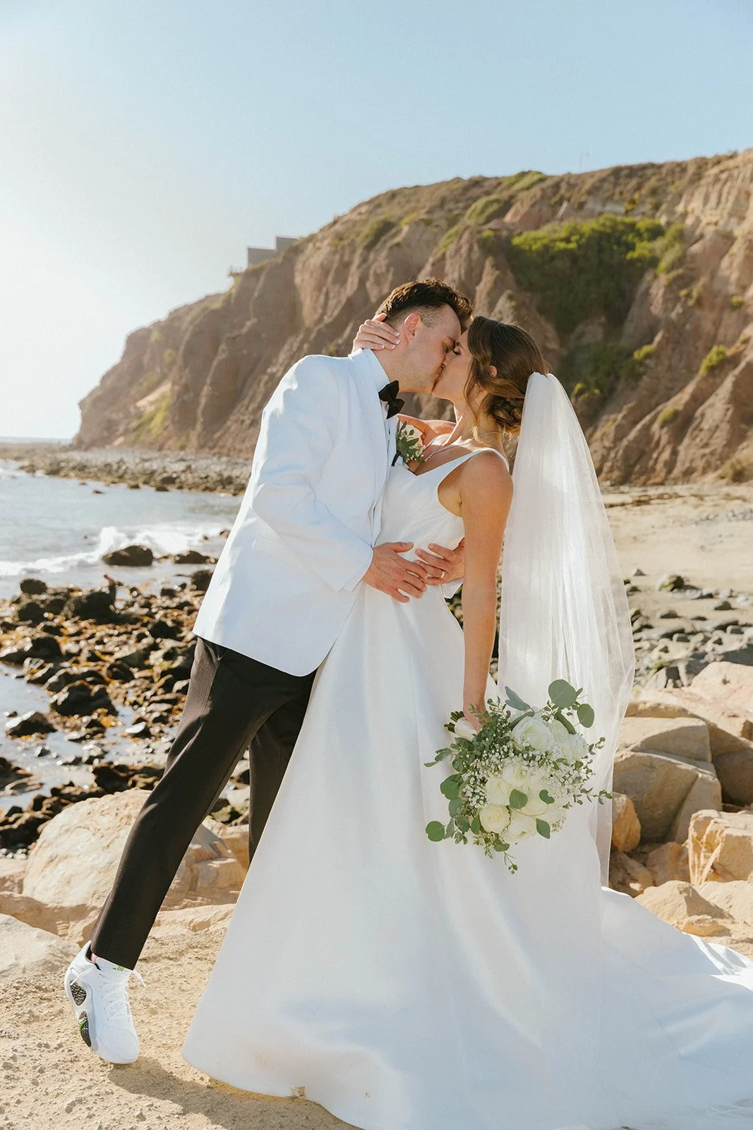 A bride and groom kissing on a rocky beach with cliffs in the background, the bride wearing a white wedding gown and veil, holding a bouquet of white flowers, and the groom in a white tuxedo jacket and black pants.