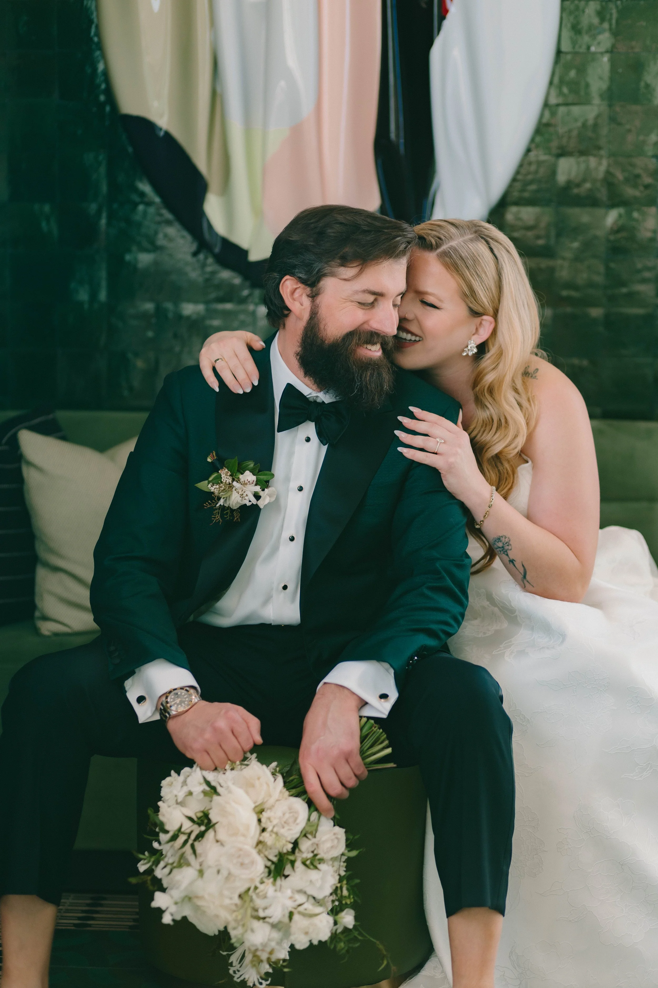 A newlywed couple sitting together, smiling and embracing, with the bride whispering into the groom's ear. The groom is dressed in a tuxedo with a boutonniere, and the bride is wearing earrings and a wedding dress. There are flowers on the table in front of them.