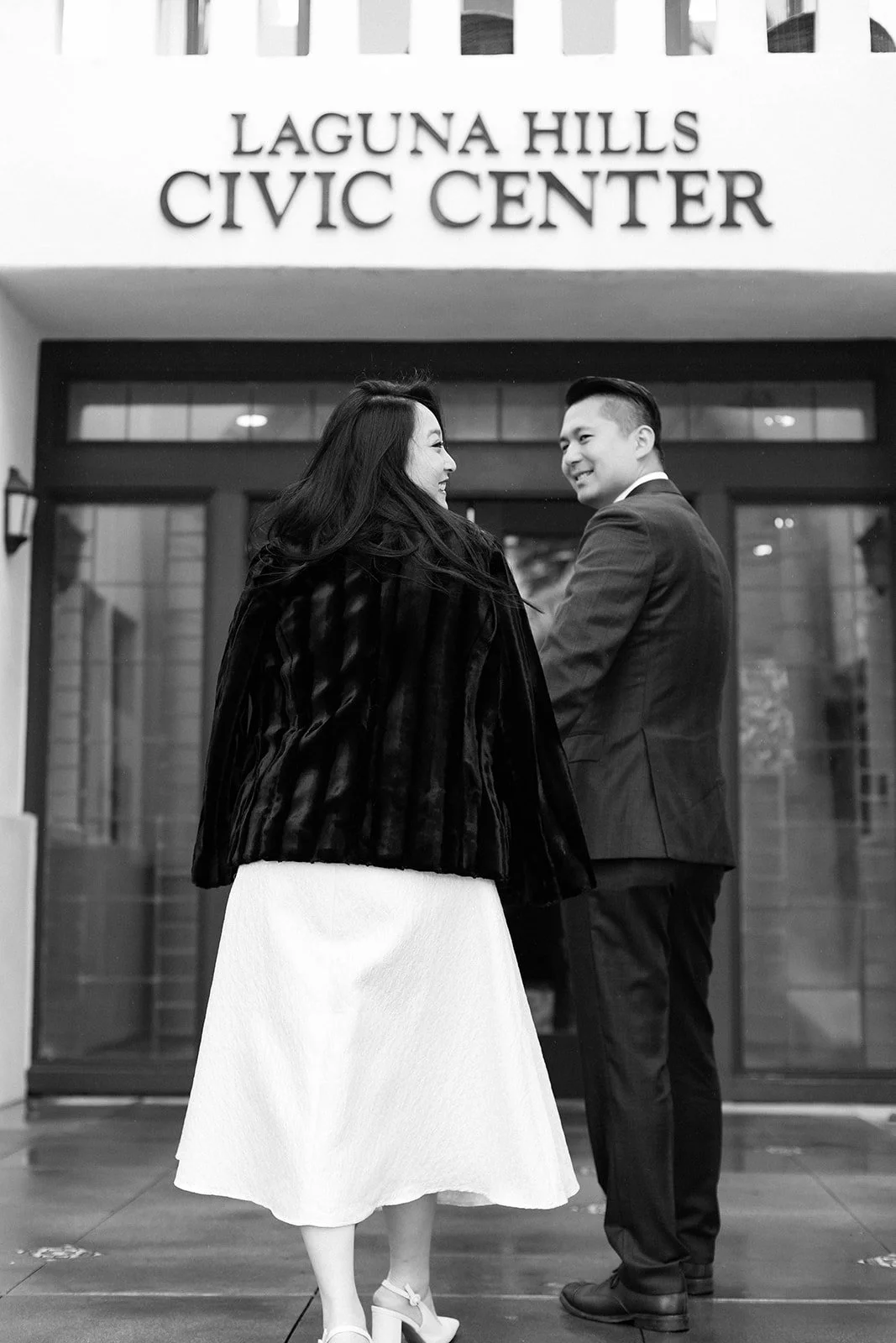 A couple dressed in formal attire standing outside the Laguna Hills Civic Center. The woman has long dark hair and is wearing a white dress with a dark fur coat, while the man is wearing a suit with a tie. They are smiling at each other.