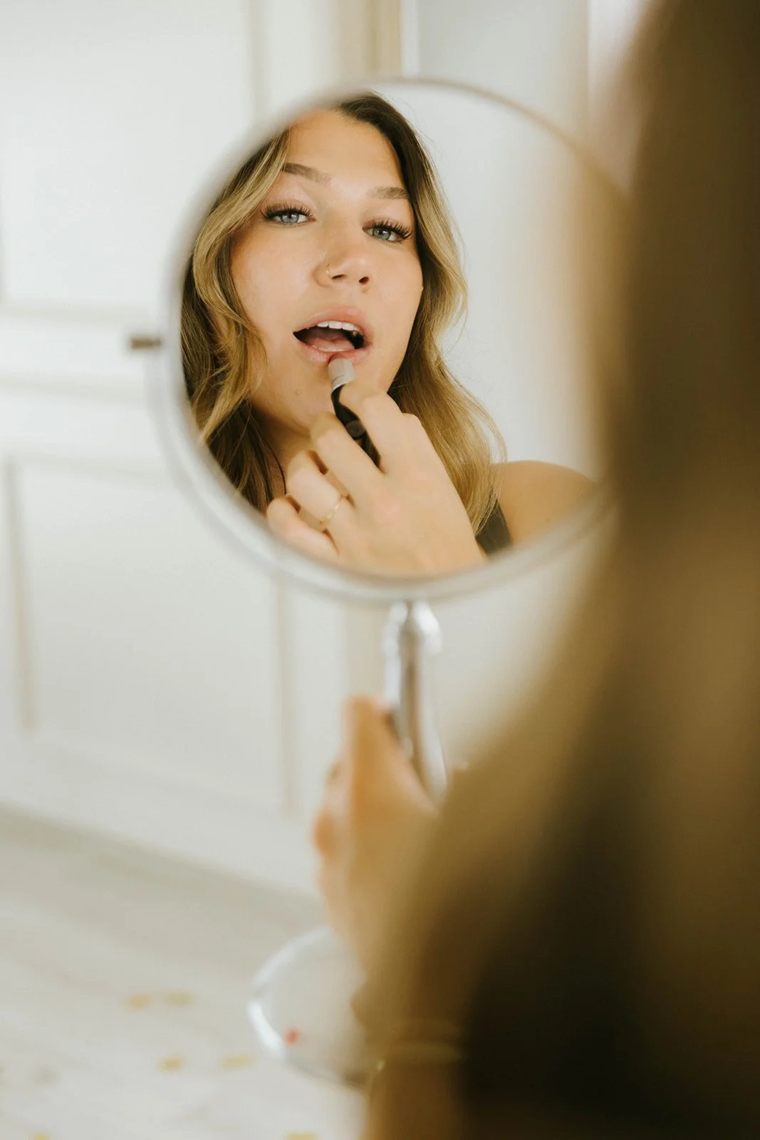 A woman applying lipstick as she looks into a round mirror.