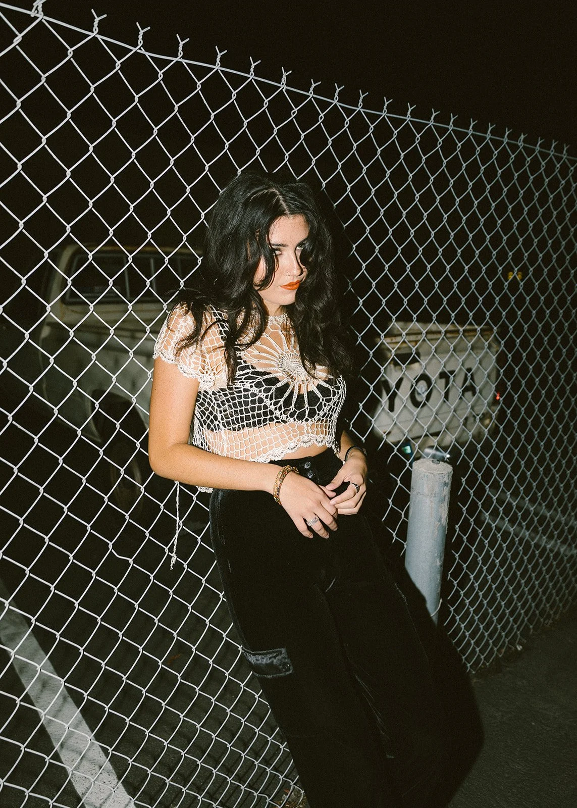 Woman with long dark hair posing in a crochet top against a chain link fence at night — Edgy fashion portrait session in Orange County — Captured by Tesla Rae Photo
