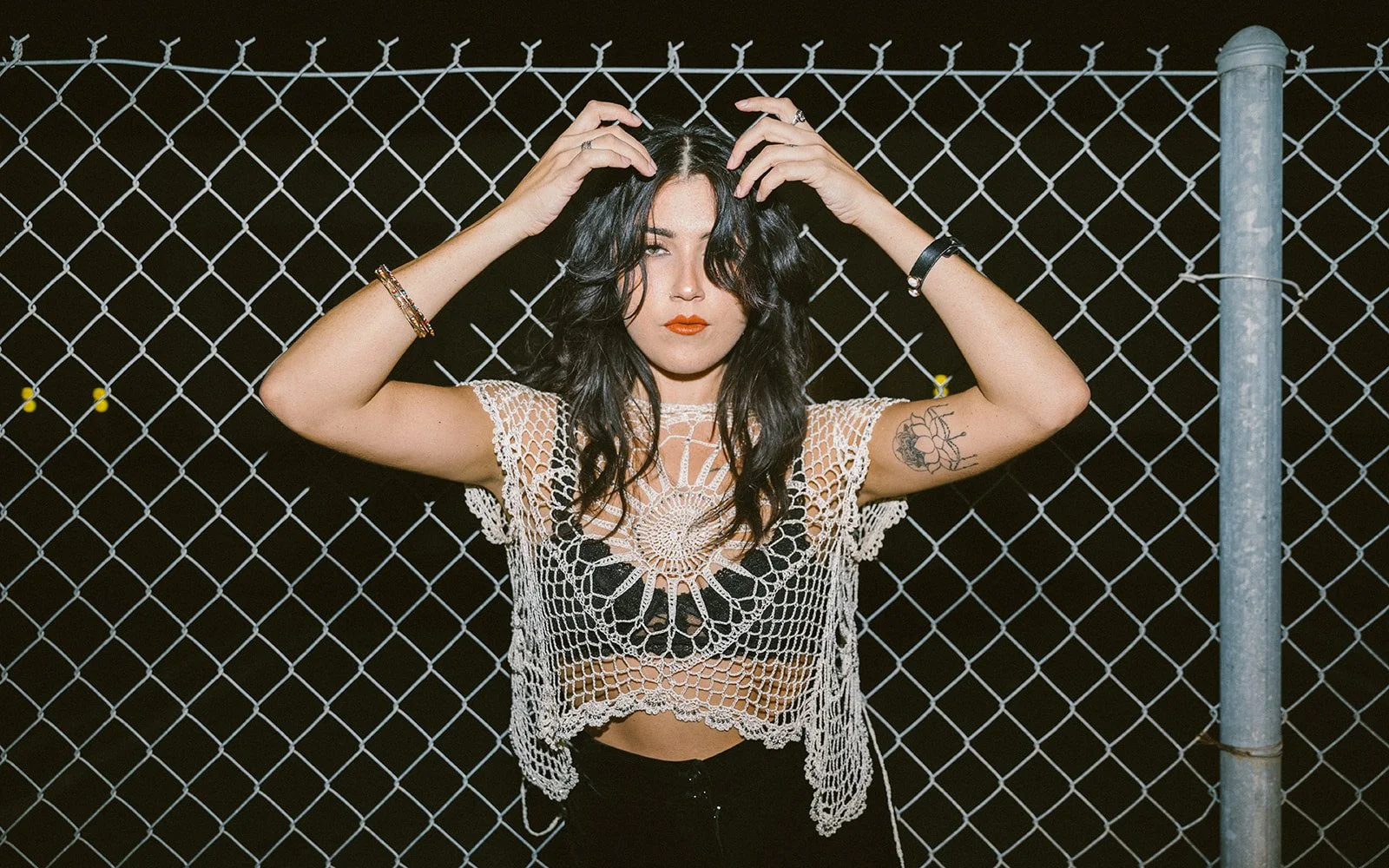Woman with long dark hair posing in a crochet top against a chain link fence at night — Edgy fashion portrait session in Orange County — Captured by Tesla Rae Photo