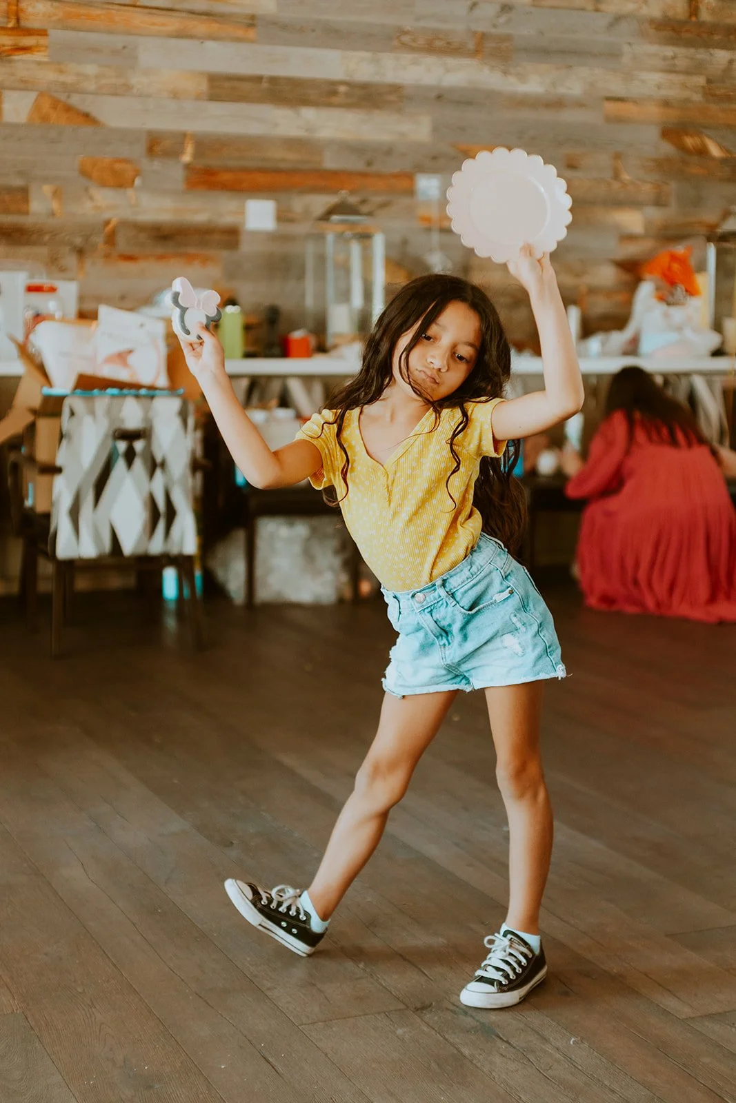 Young girl in a yellow shirt and denim shorts dancing in a room with a wooden wall, holding a paper plate and a small object.