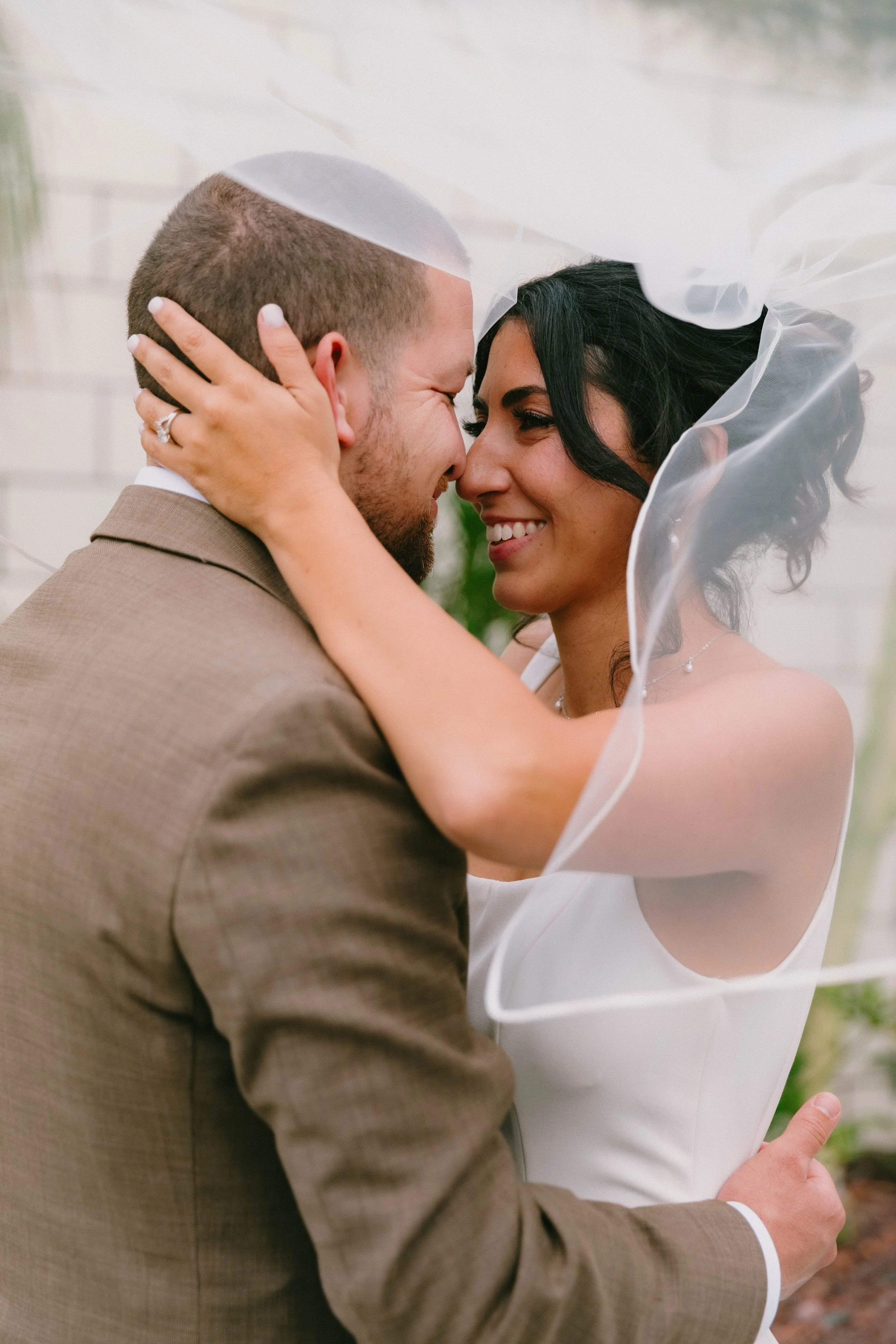 A close-up of a newly married couple embracing each other, smiling with eyes closed, with a veil draped over them.