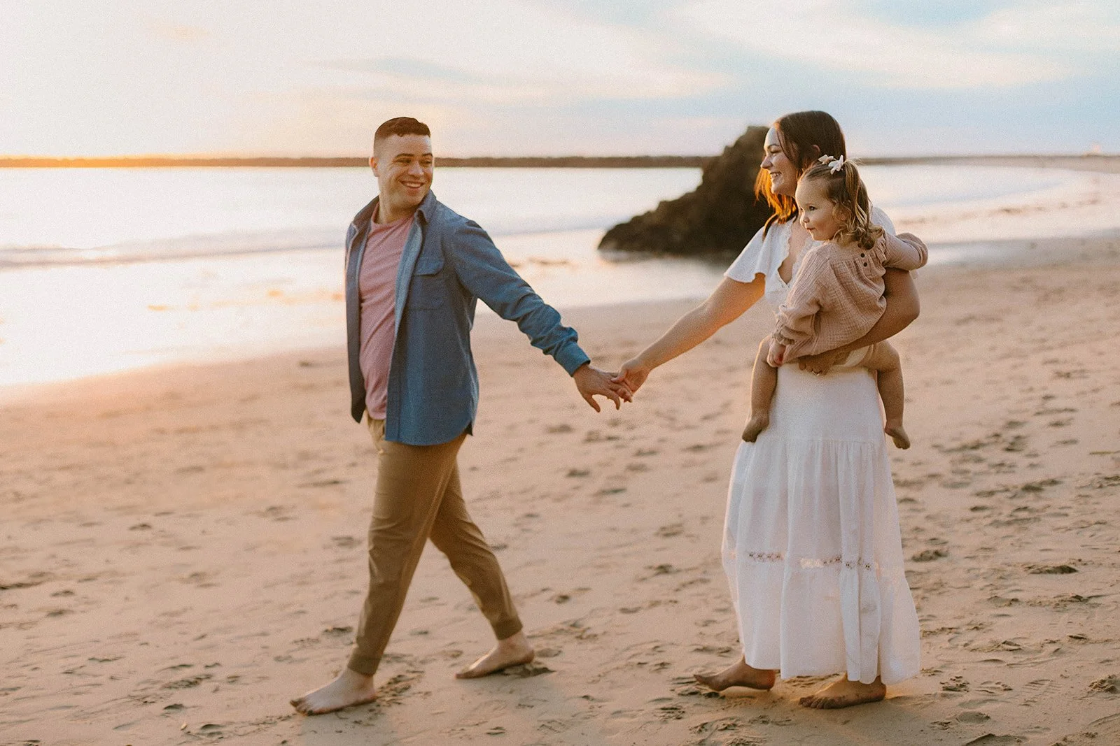 A family of three holding hands and walking on a beach at sunset. The mother, carrying her young daughter, and the father are smiling and enjoying the moment.