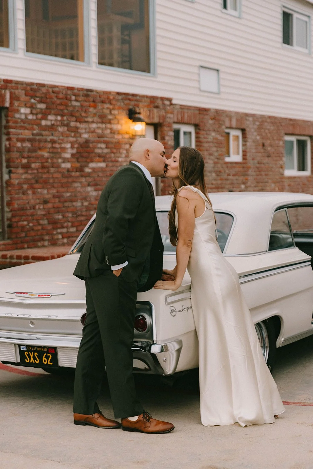 A bride and groom sharing a kiss beside a vintage white car outside a coastal venue at sunset. Elegant and cinematic wedding portrait with a romantic editorial feel.