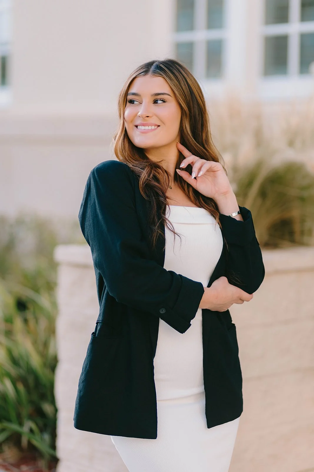 A young woman with long brown hair looking to her left, smiling, wearing a black blazer over a white dress, standing outdoors near a building with large windows and some greenery in the background.