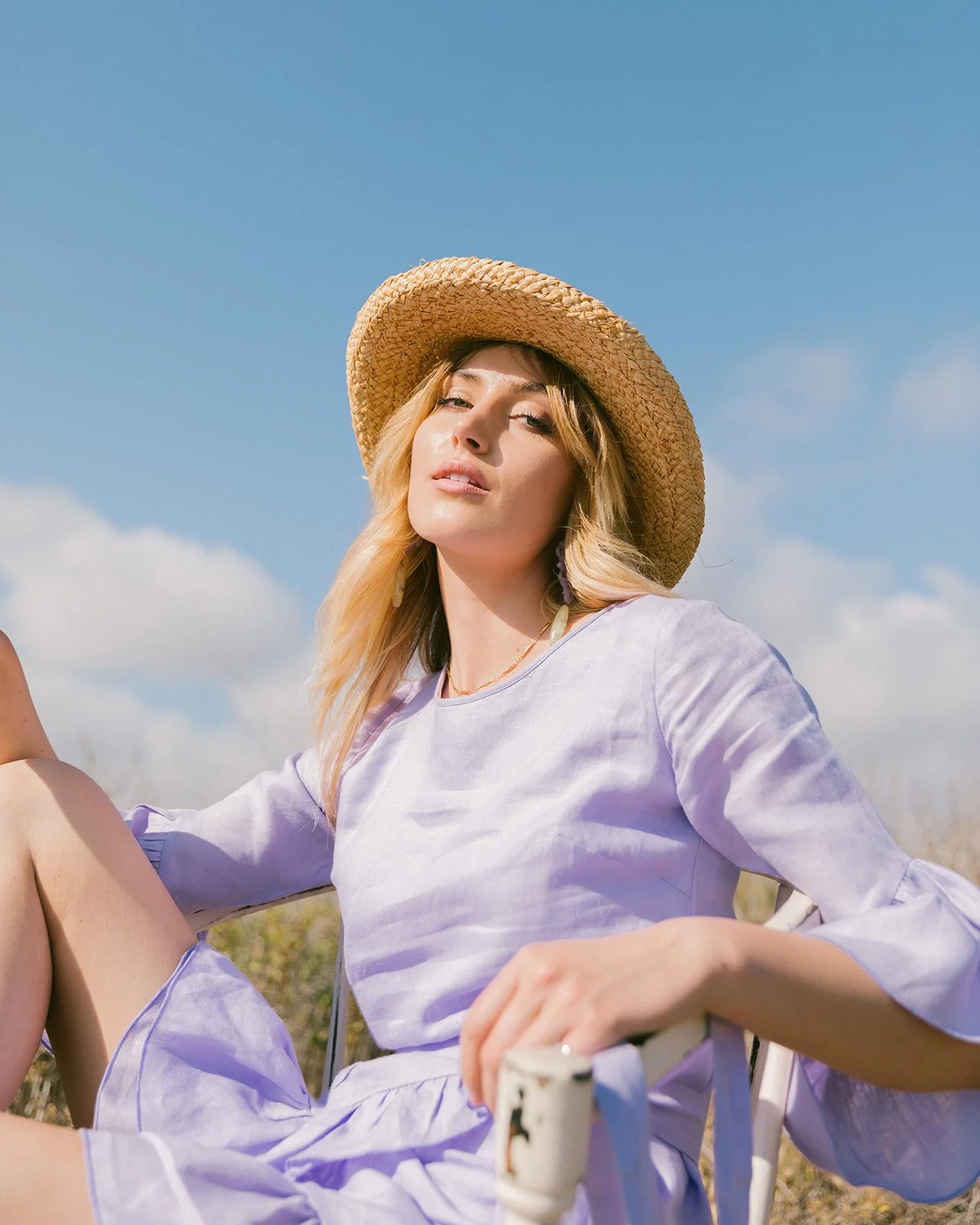 A woman with blonde hair wearing a straw hat and a light purple outfit, sitting outdoors on a sunny day with blue sky and clouds in the background.