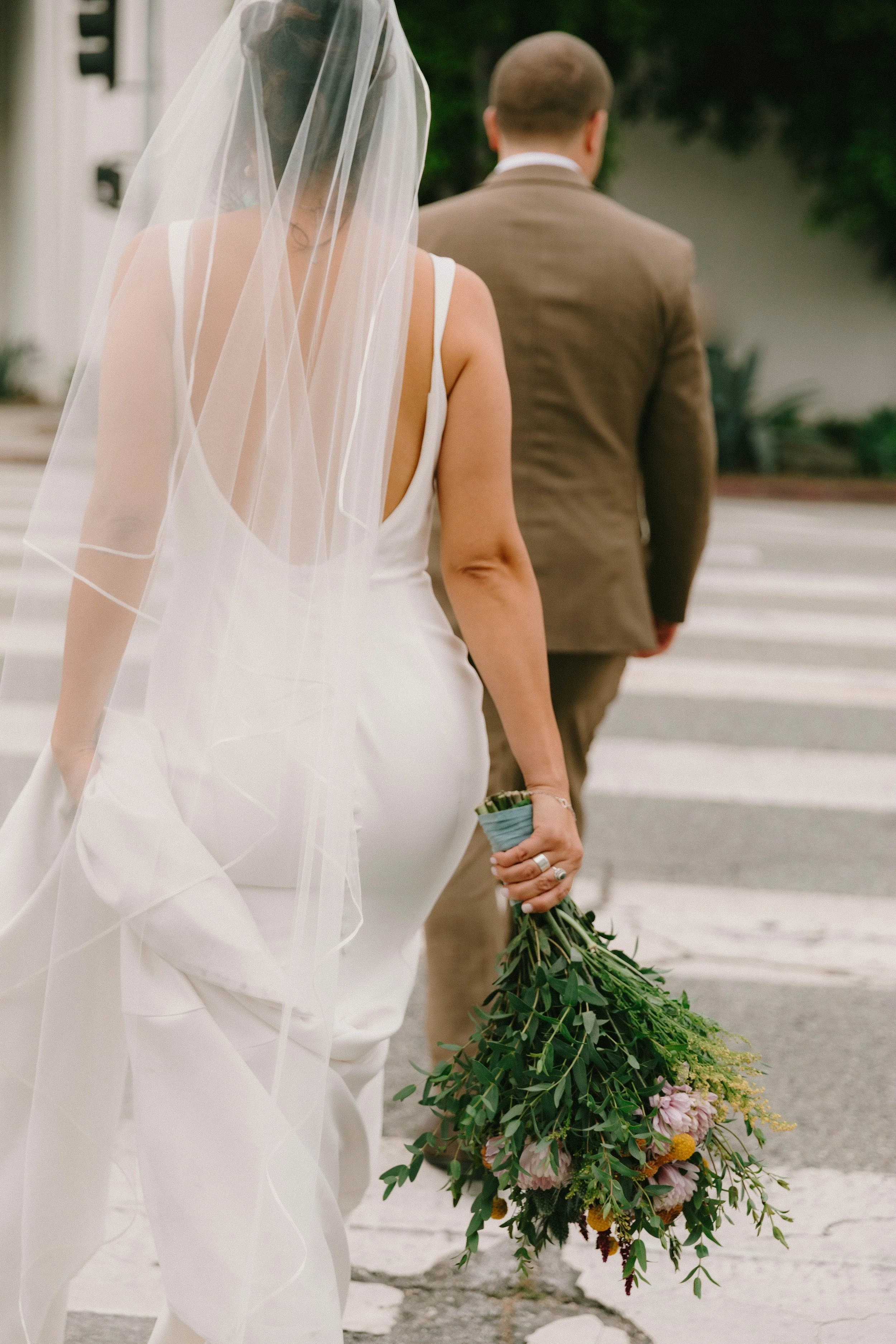 A bride in a white wedding dress and veil holding a bouquet of flowers, walking with a groom dressed in a tan suit, holding a large bouquet of greenery and flowers.