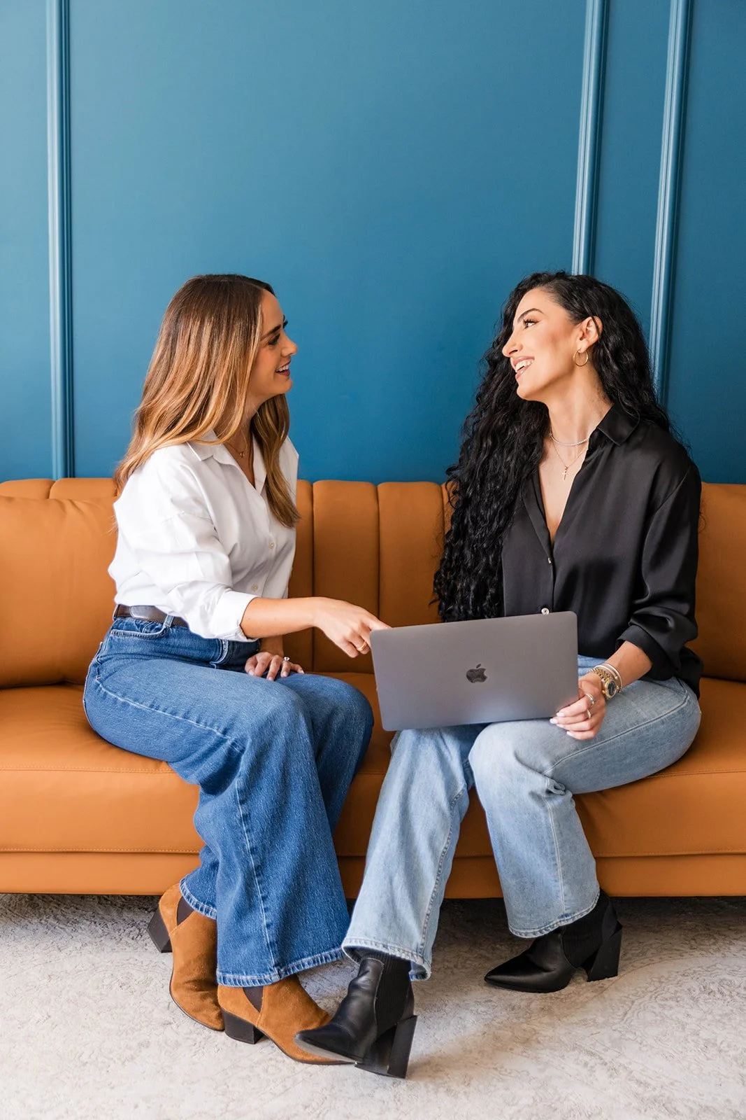 Two women are sitting on a tan couch, engaged in a friendly conversation. One woman is pointing at a laptop, while the other is smiling. The background is a blue wall.