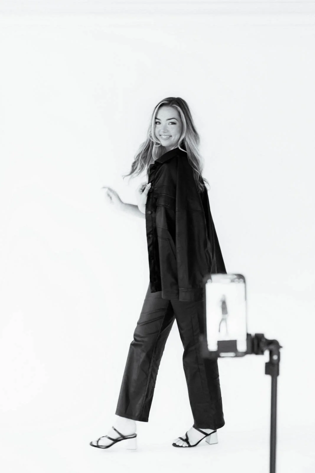 A woman with long hair smiling and posing in front of a white background, wearing a leather jacket, wide-leg pants, and high heels, as a phone on a tripod records her.