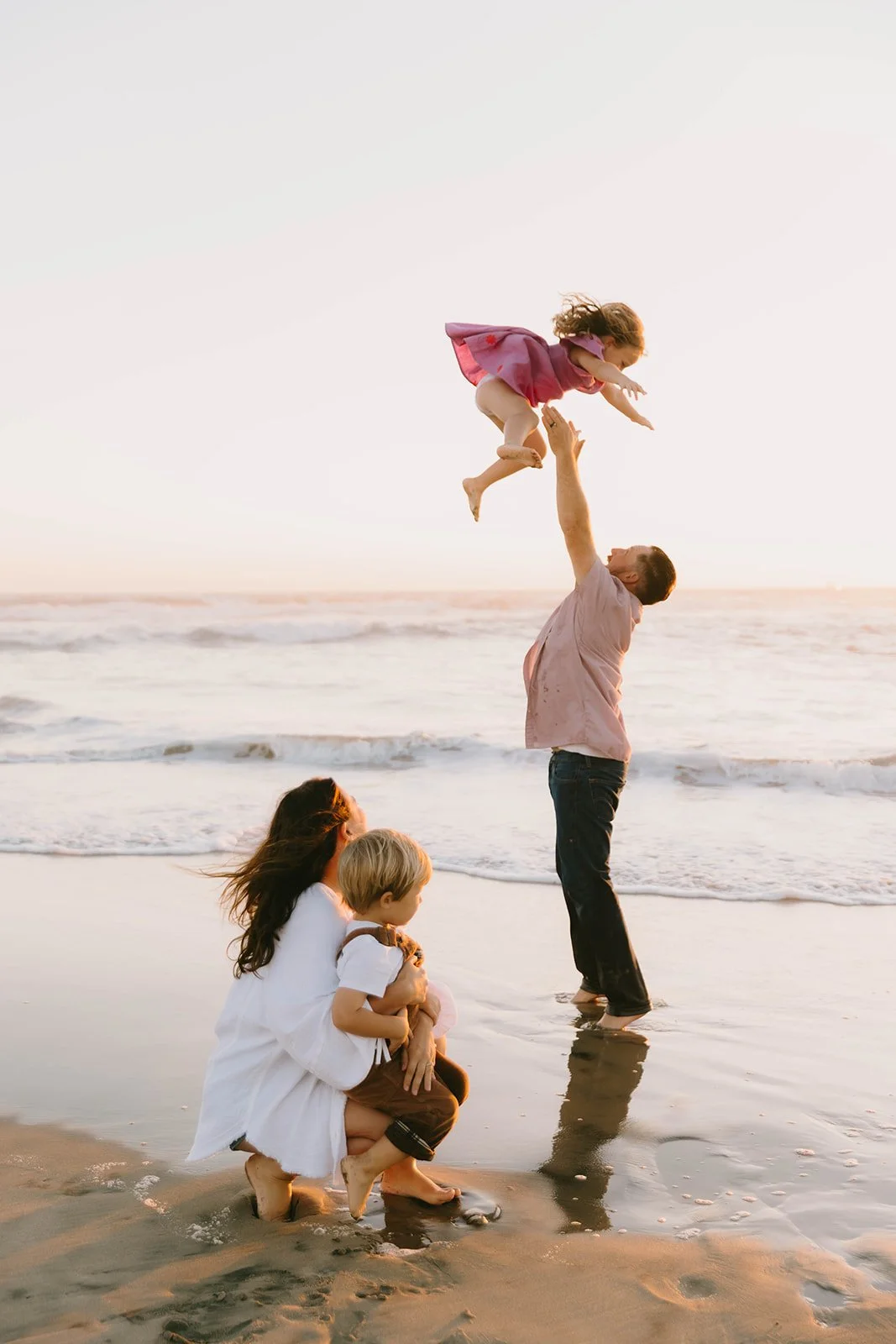 A father tossing his young daughter into the air while the family enjoys a golden hour beach session in Huntington Beach, California. Candid coastal family photography with warm sunset light and playful natural moments.