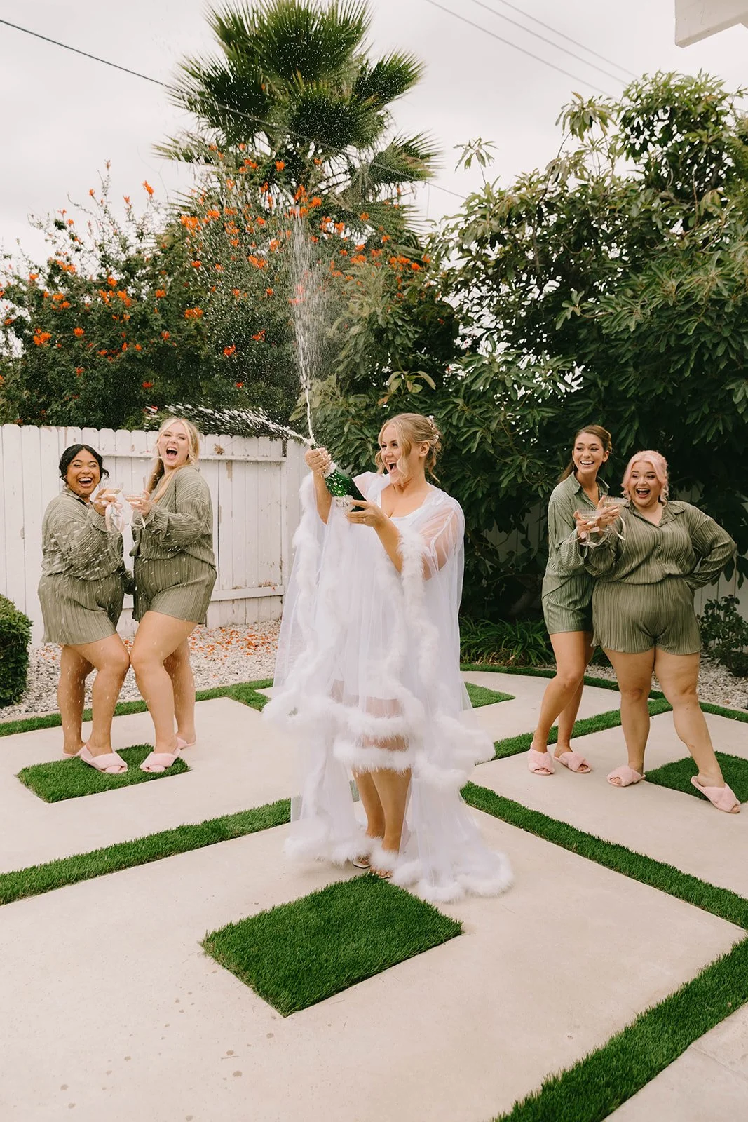 A bride in a sheer white getting-ready robe celebrating with her bridesmaids during a champagne spray moment outdoors. Fun candid wedding morning photography with champagne coupes, matching bridesmaid outfits, and joyful energy.