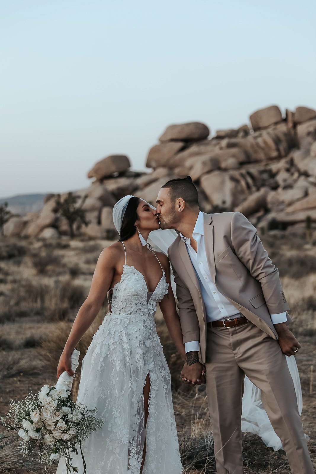 A newlywed couple sharing a kiss in a desert landscape with Joshua trees and mountains in the background. Destination wedding with Elopement photographer.