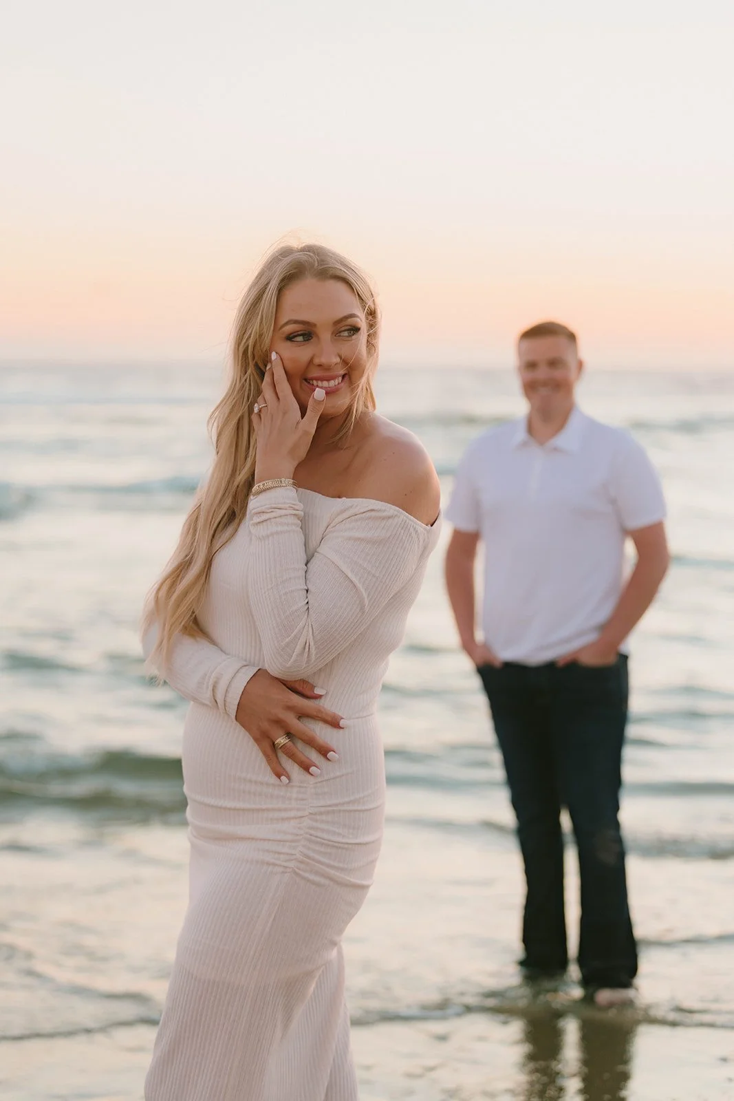 A newly engaged couple standing together in the ocean at sunset, with the woman smiling toward the camera while showing her engagement ring. Romantic beach engagement photos in Orange County with soft pastel sunset tones.