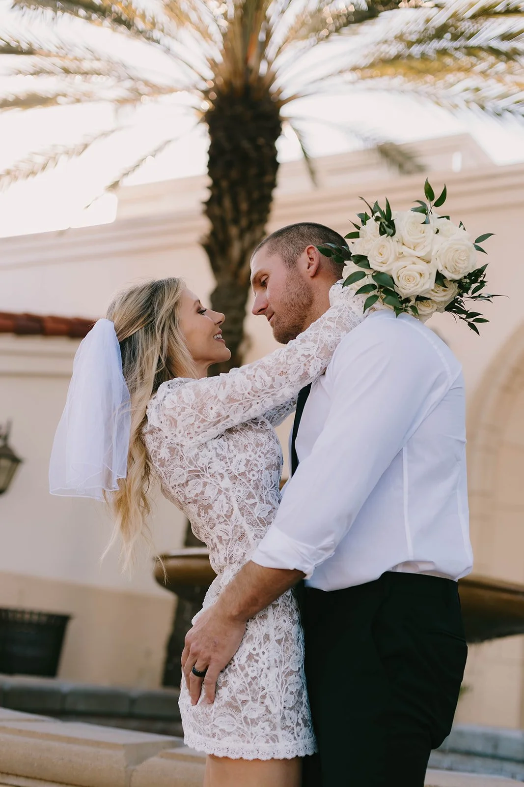 A groom dipping his bride backward beneath tall palm trees outside a Southern California courthouse, her veil flowing behind her as they smile together in the sunlight. Romantic courthouse elopement portrait with an elegant coastal California feel.