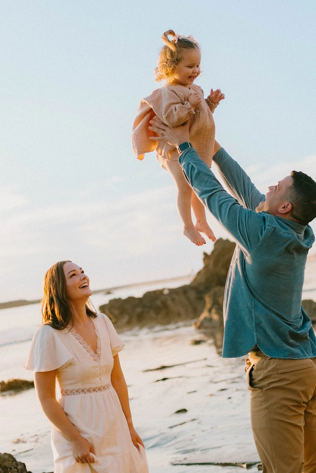 A candid Orange County beach family session at golden hour featuring a father lifting his daughter in the air while mom smiles nearby along the shoreline. Warm coastal family photography with playful movement and emotional connection.