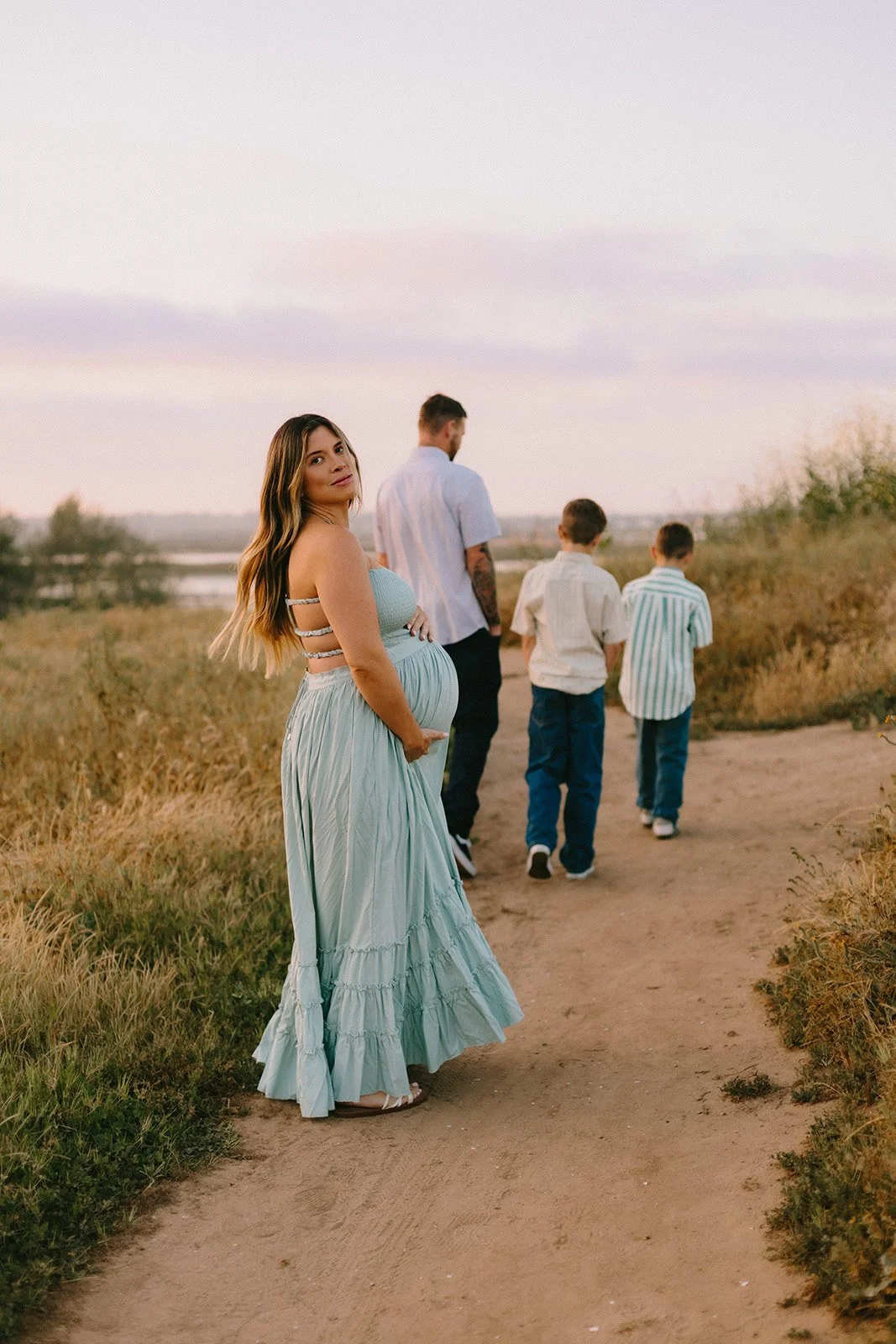 A pregnant woman in a light blue dress standing on a dirt path with her family, including a man and three boys, in a natural outdoor setting during sunset.