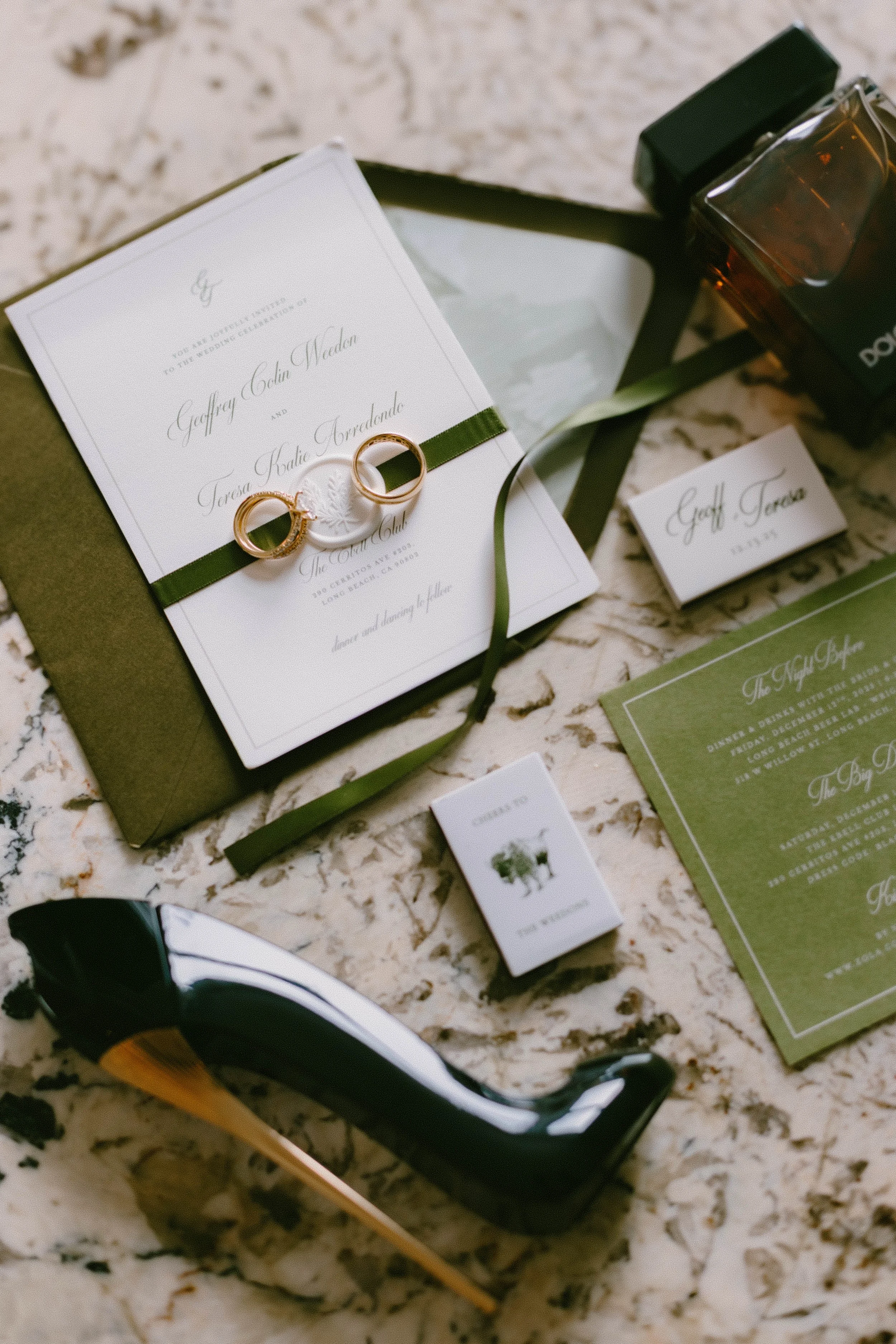 Wedding invitation with two gold rings, perfume bottles, sunglasses, and various cards on a marble surface.