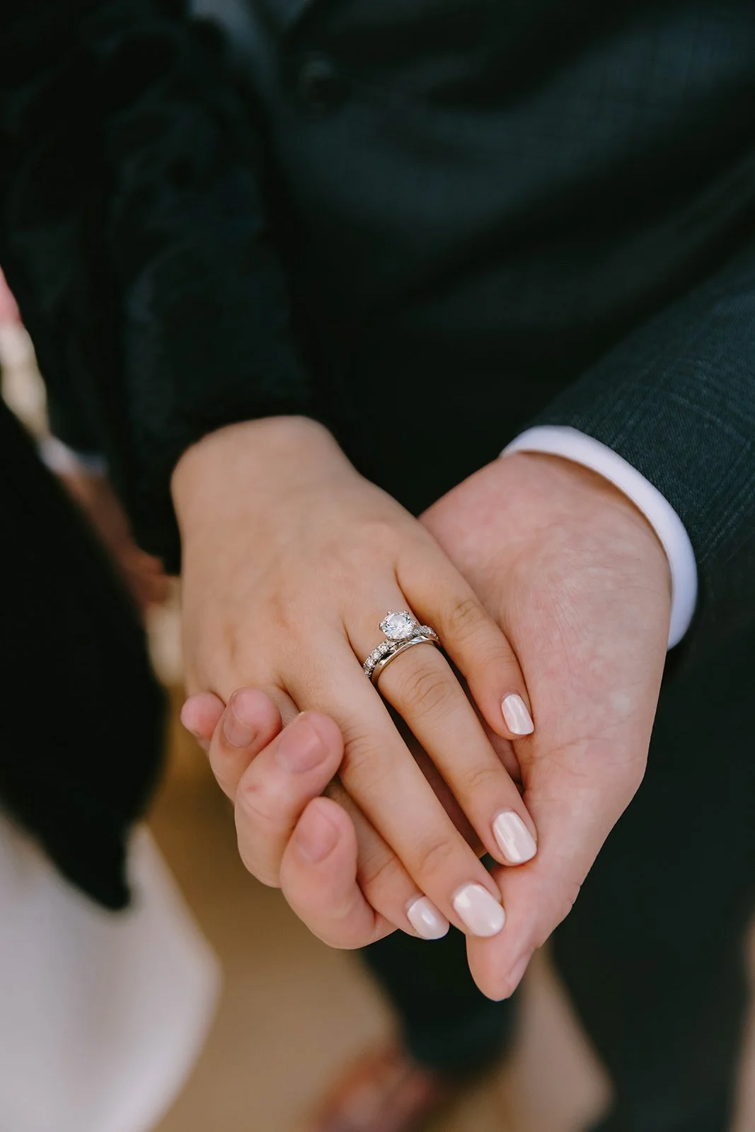 Close-up of a hand wearing a diamond engagement ring and wedding band, held by another hand, with a person in a dark suit in the background.