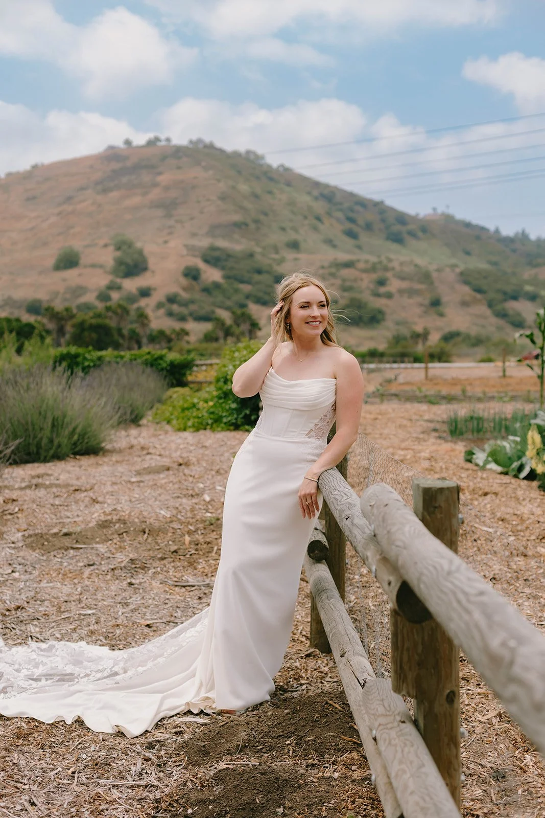 A bride in a white wedding dress standing outdoors beside a wooden fence with a hill and cloudy sky in the background.