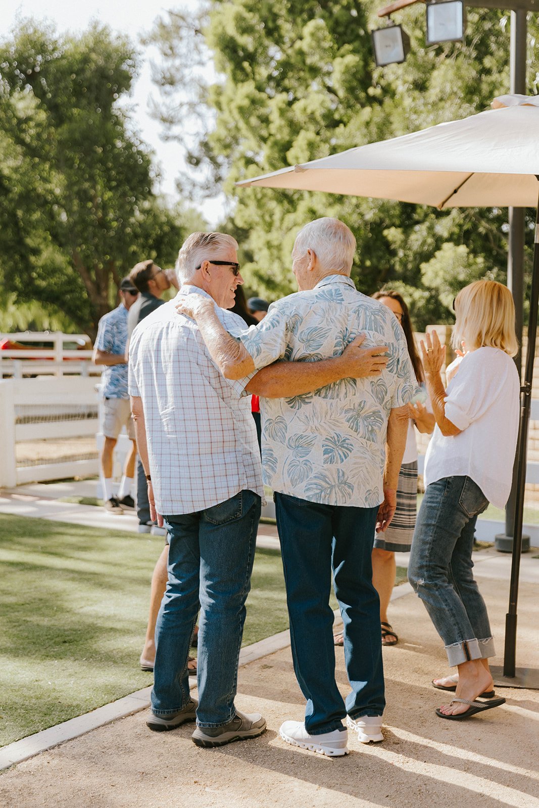 A group of people standing outdoors, laughing and talking under a large umbrella, with trees in the background.