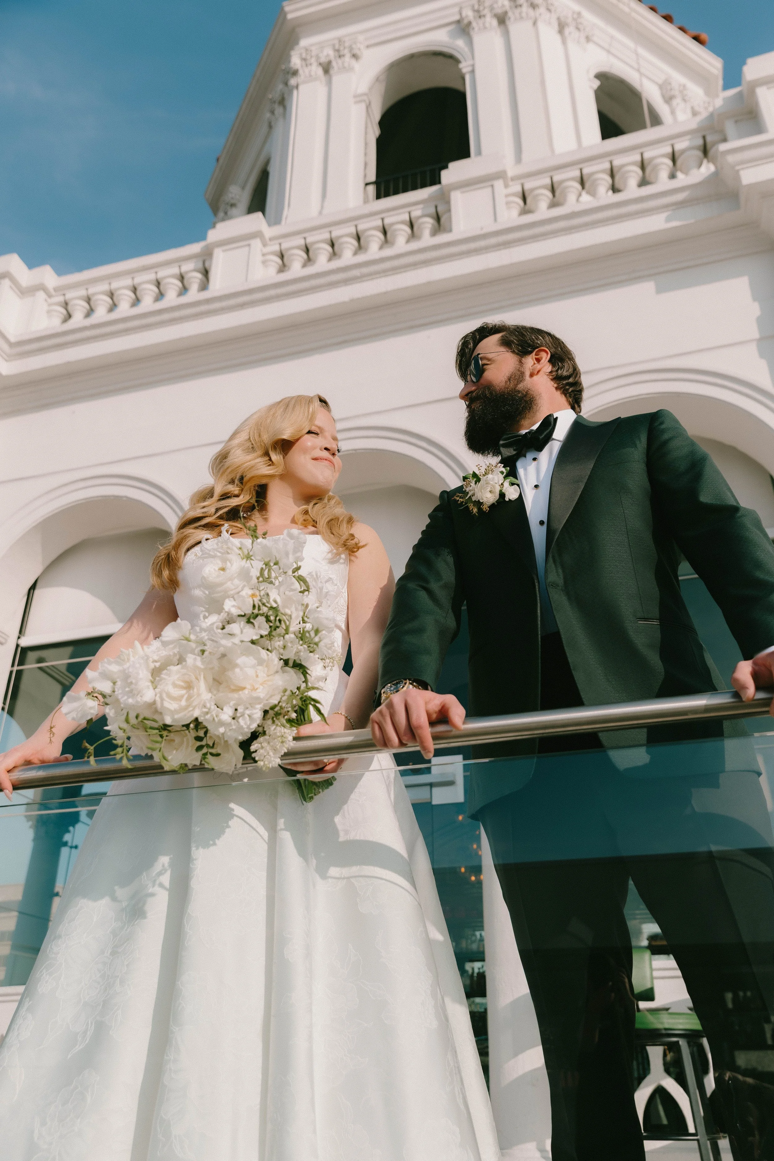 A newlywed couple embracing on a grand balcony overlooking a luxury hotel courtyard at sunset. Editorial wedding portrait with romantic architecture, warm light, and an elegant cinematic feel.