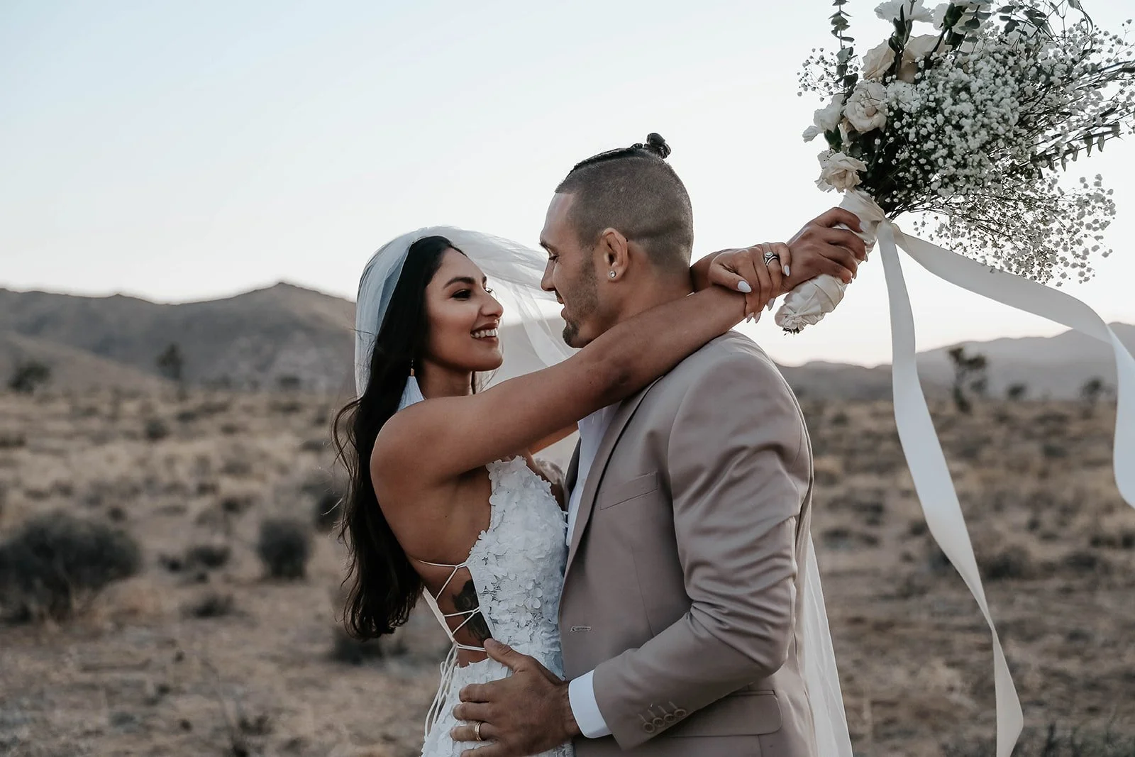 A newlywed couple sharing a kiss in a desert landscape with Joshua trees and mountains in the background. Destination wedding with Elopement photographer.