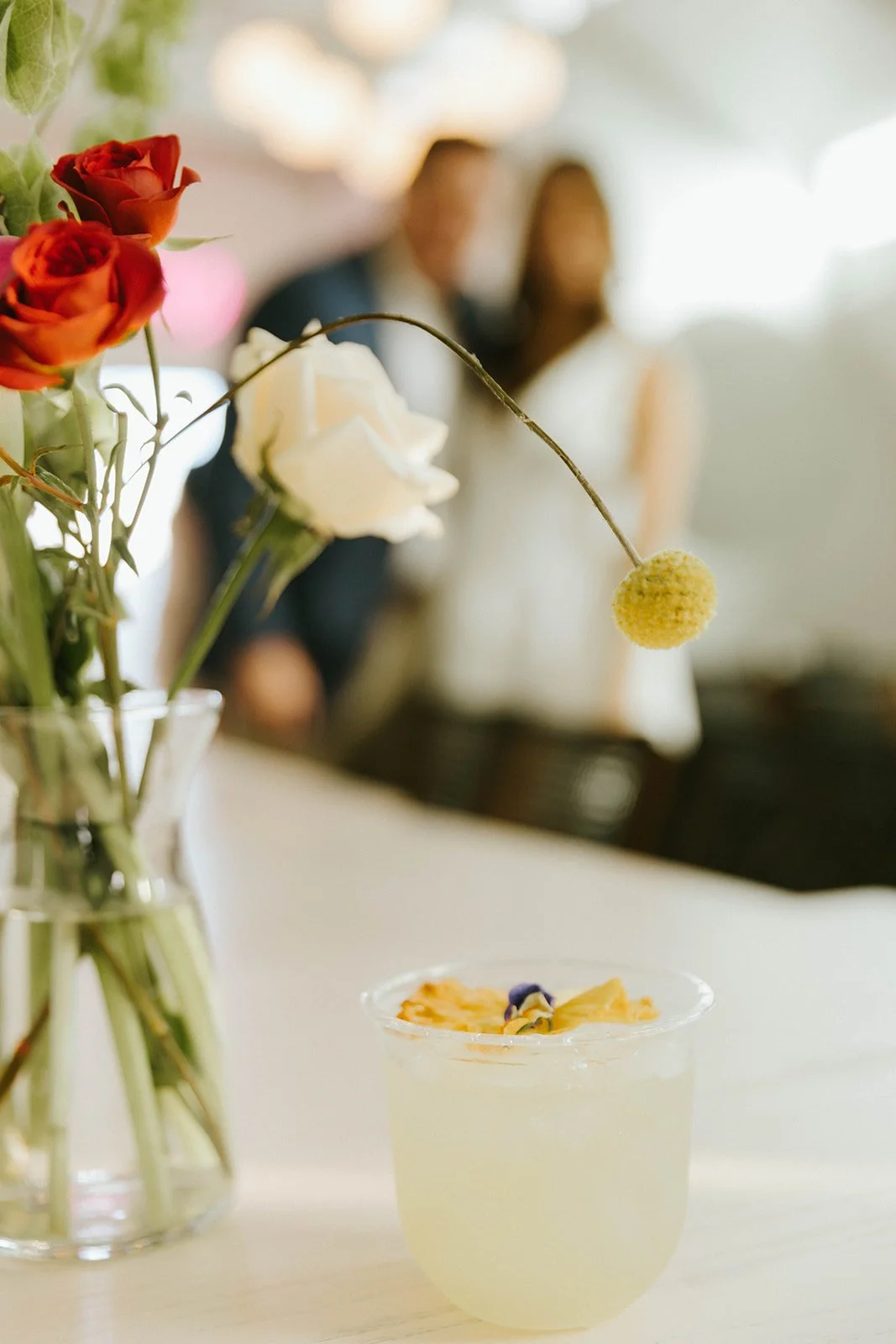 A clear glass vase with red and white flowers on a white table. In the foreground is a yellow cocktail with a flower garnish, and in the background are blurred people.