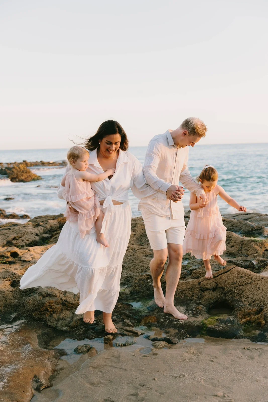 Candid Laguna Beach family photography session at golden hour featuring a family walking barefoot along the coastal rocks in soft neutral tones. Emotional Orange County family portraits with natural movement and connection.