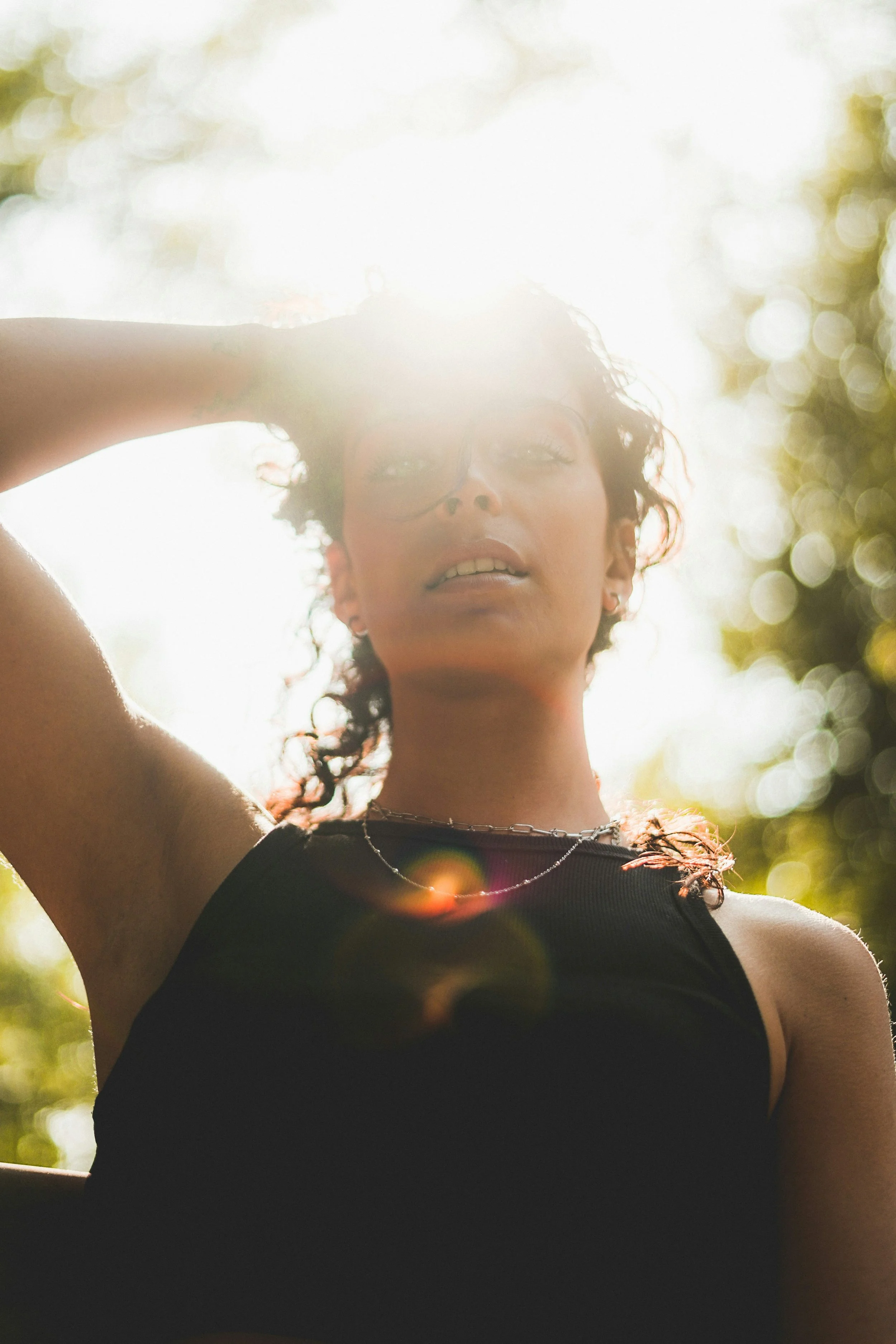 A woman with curly hair and hoop earrings, wearing a black sleeveless top and a necklace, standing outdoors with sunlight filtering through trees, creating a bokeh effect in the background.