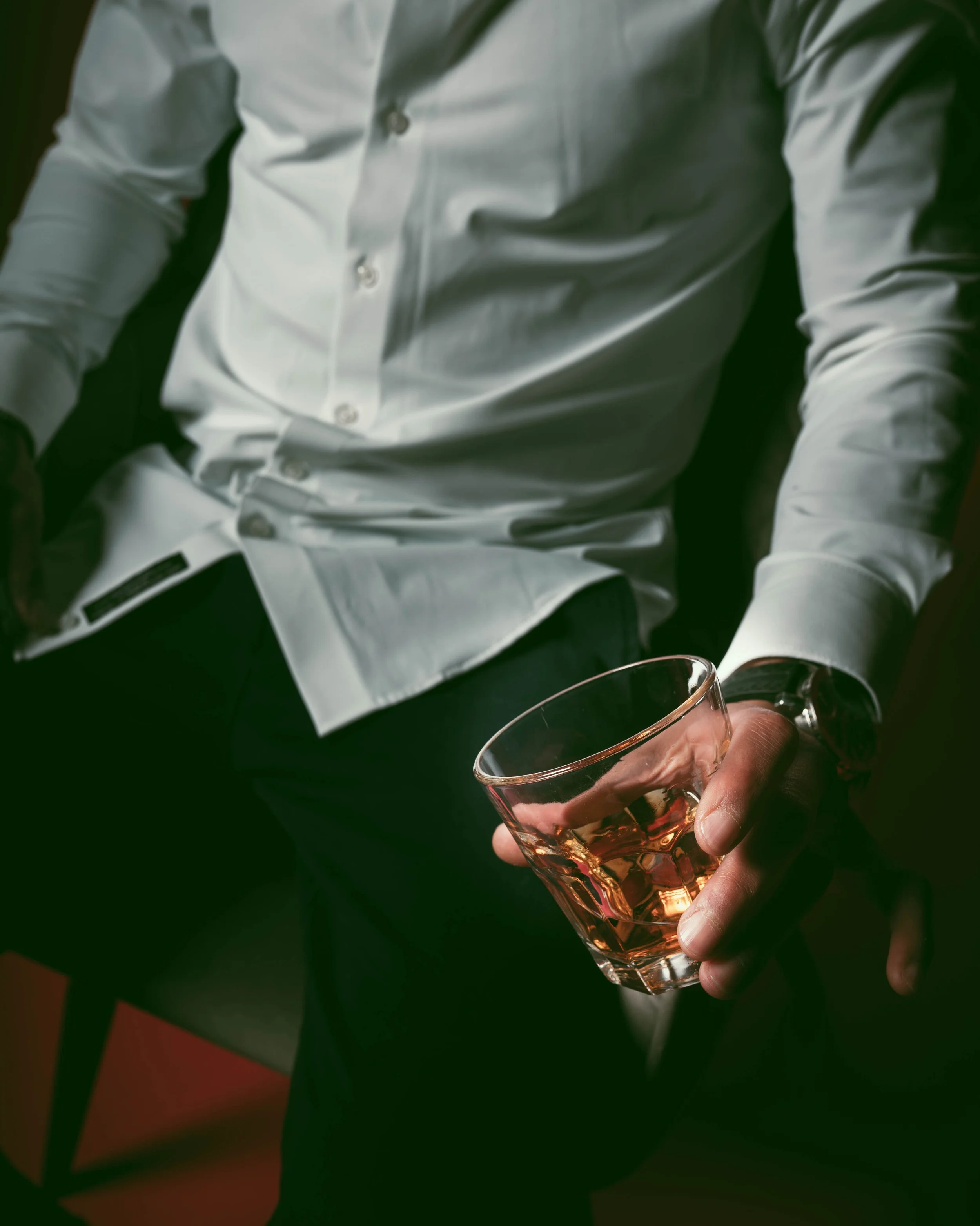 A man in a white dress shirt sitting and holding a glass of whiskey.