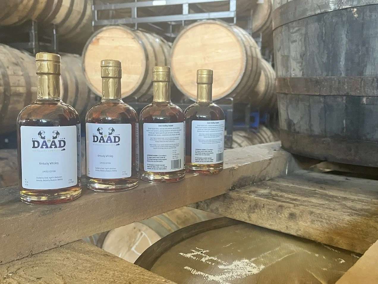 Four bottles of Daad Kentucky Whiskey lined up on a wooden shelf in a distillery with wooden barrels in the background.
