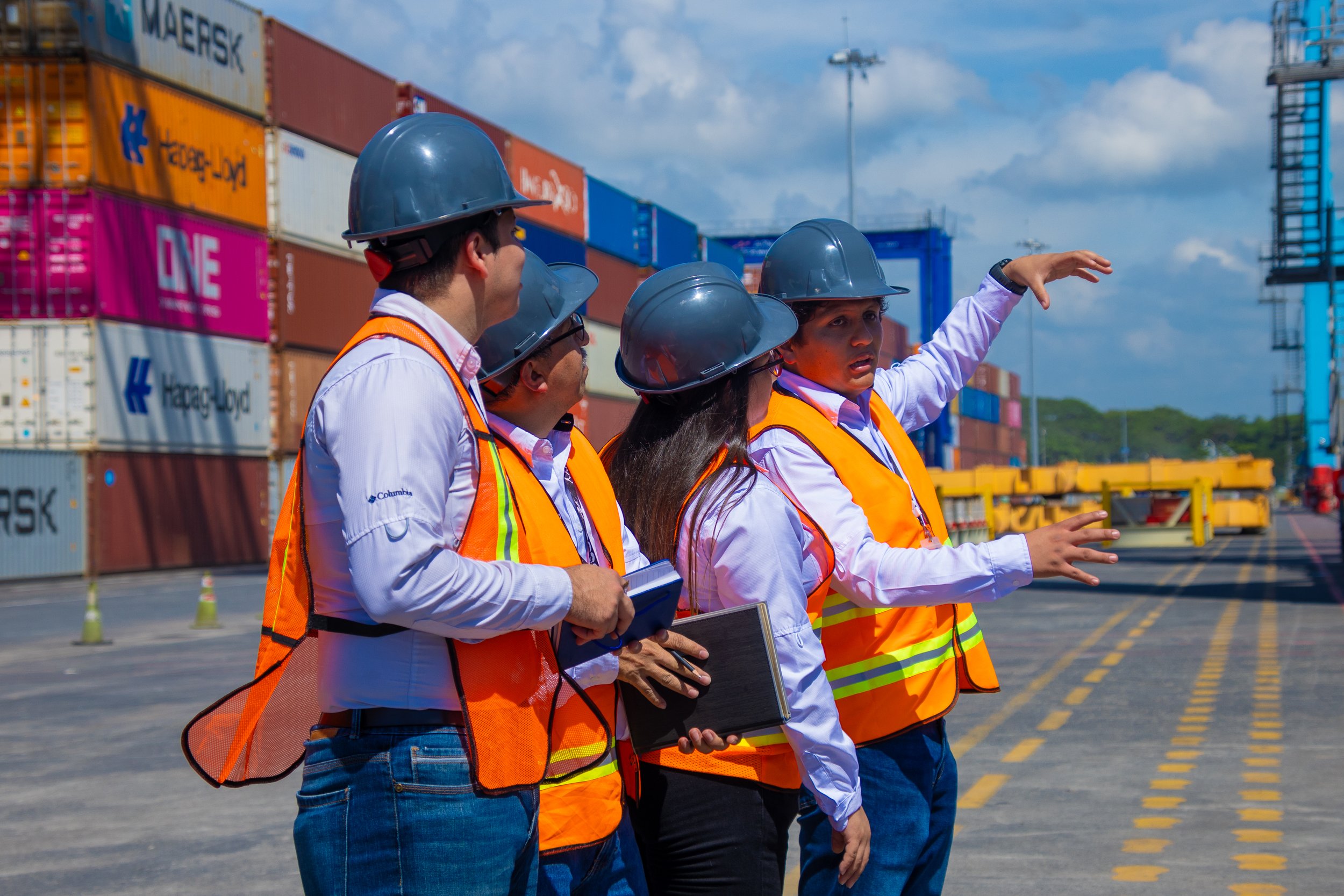 Cuatro personas con cascos de seguridad y chalecos reflectantes discutiendo en un terminal de contenedores con montones de cajas y maquinaria de carga en el fondo.