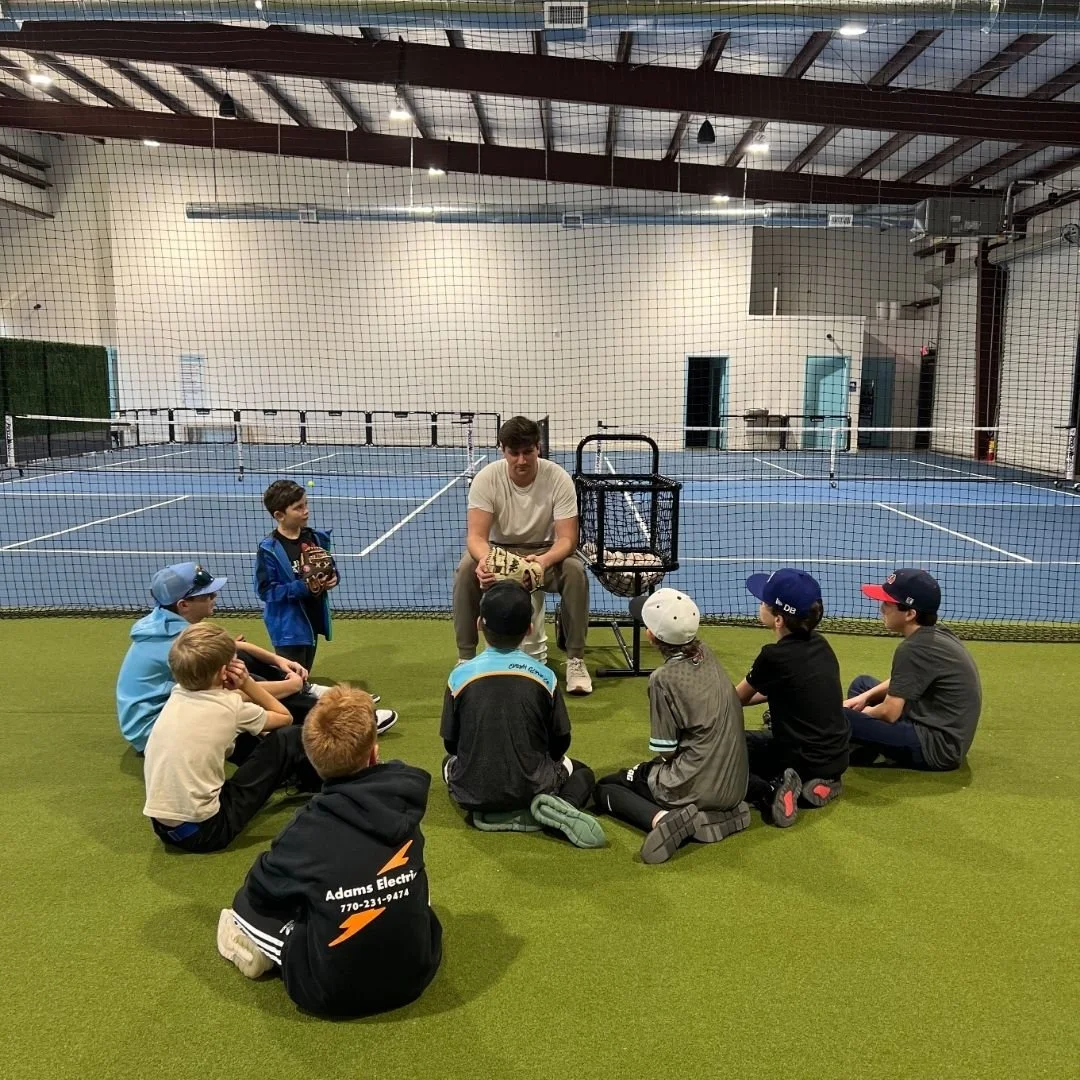 A man giving a tennis lesson to a group of children sitting on artificial grass in an indoor tennis facility, with tennis courts in the background.