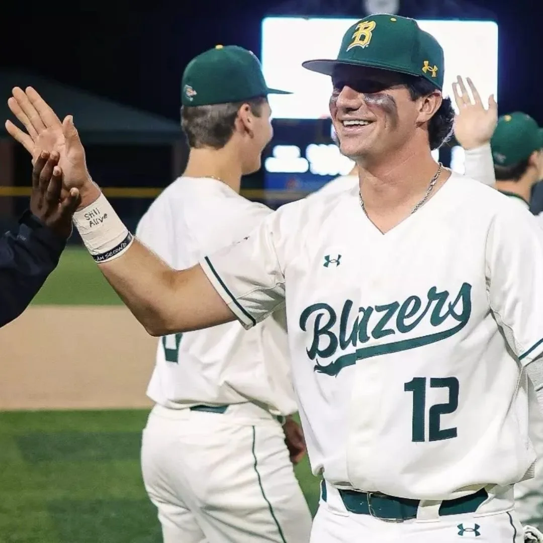 A baseball player in a white uniform and a green cap is smiling and giving a high five during a celebration. His uniform has the word 'Buzzers' and the number 12. Other players are in the background on the field.