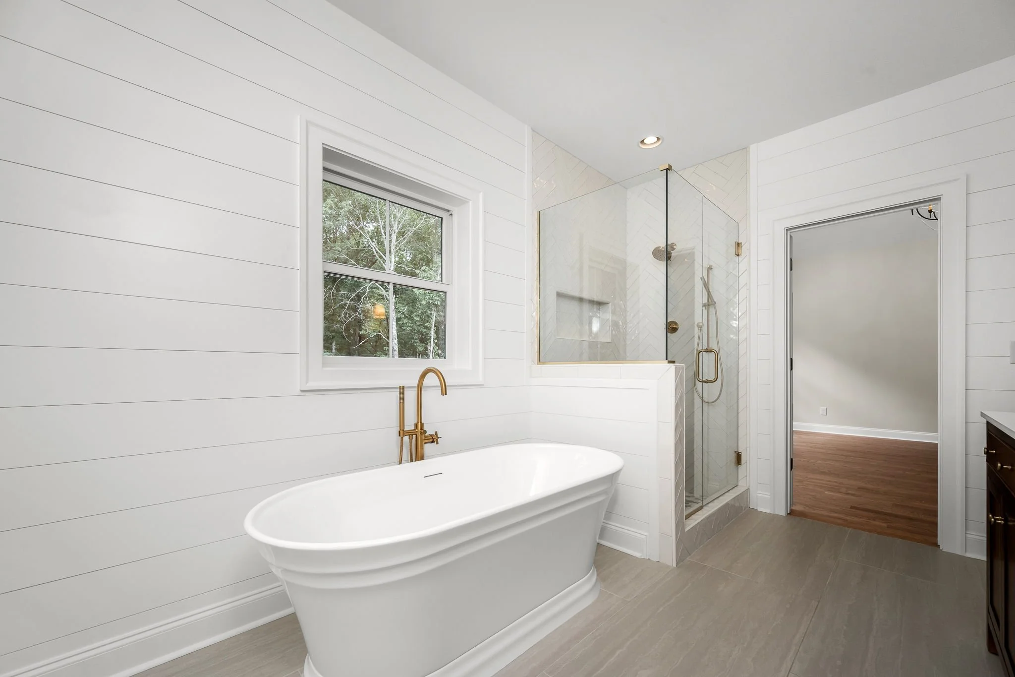 A modern bathroom with a white freestanding bathtub, a large window showing trees outside, a glass shower enclosure with gold fixtures, and wood flooring leading to a doorway.