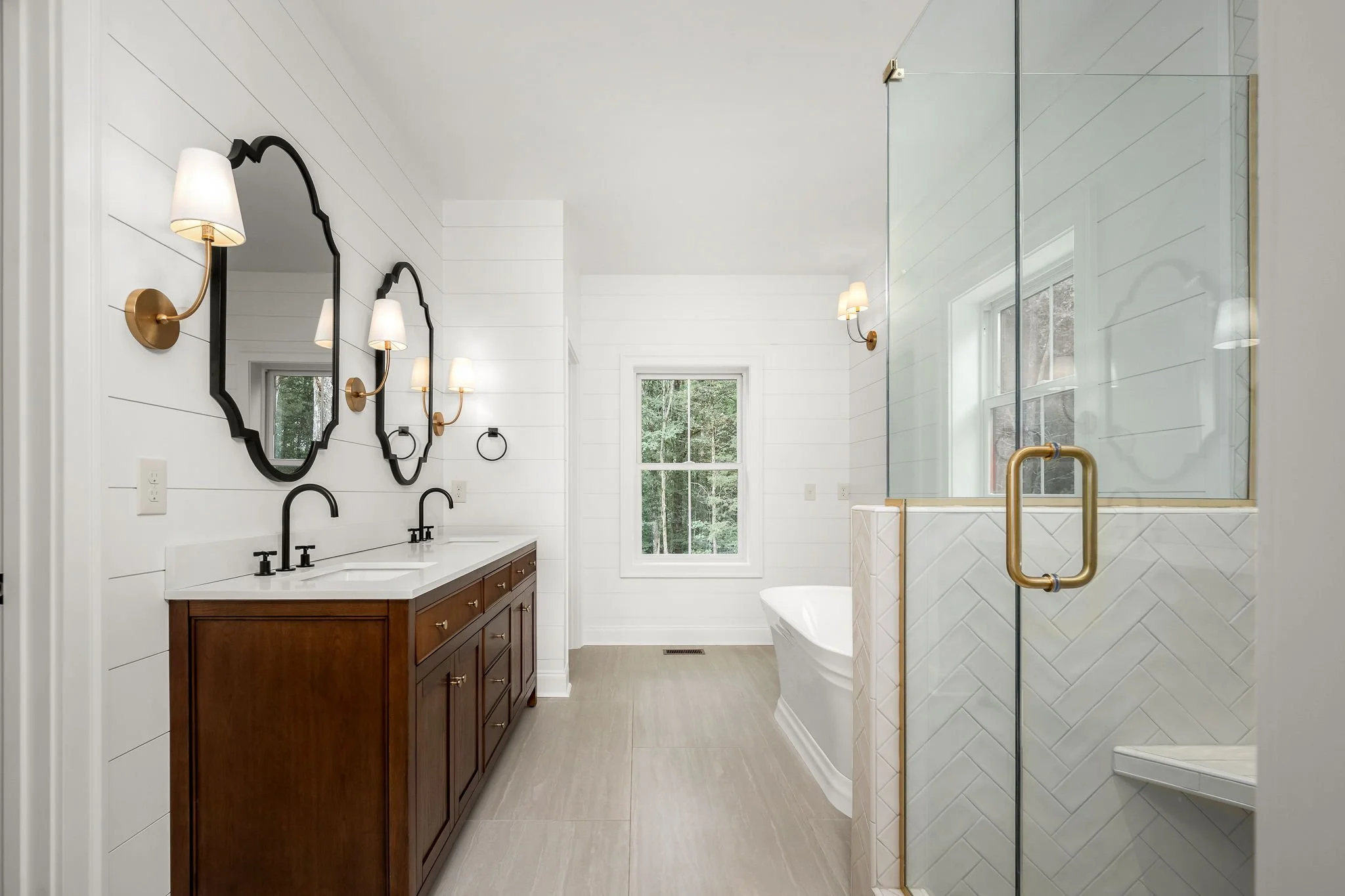 Modern bathroom with white shiplap walls, a large window, and a glass shower door with gold accents. A wooden vanity with a white countertop and two black faucets is visible, along with three wall-mounted light fixtures with white shades and black fr