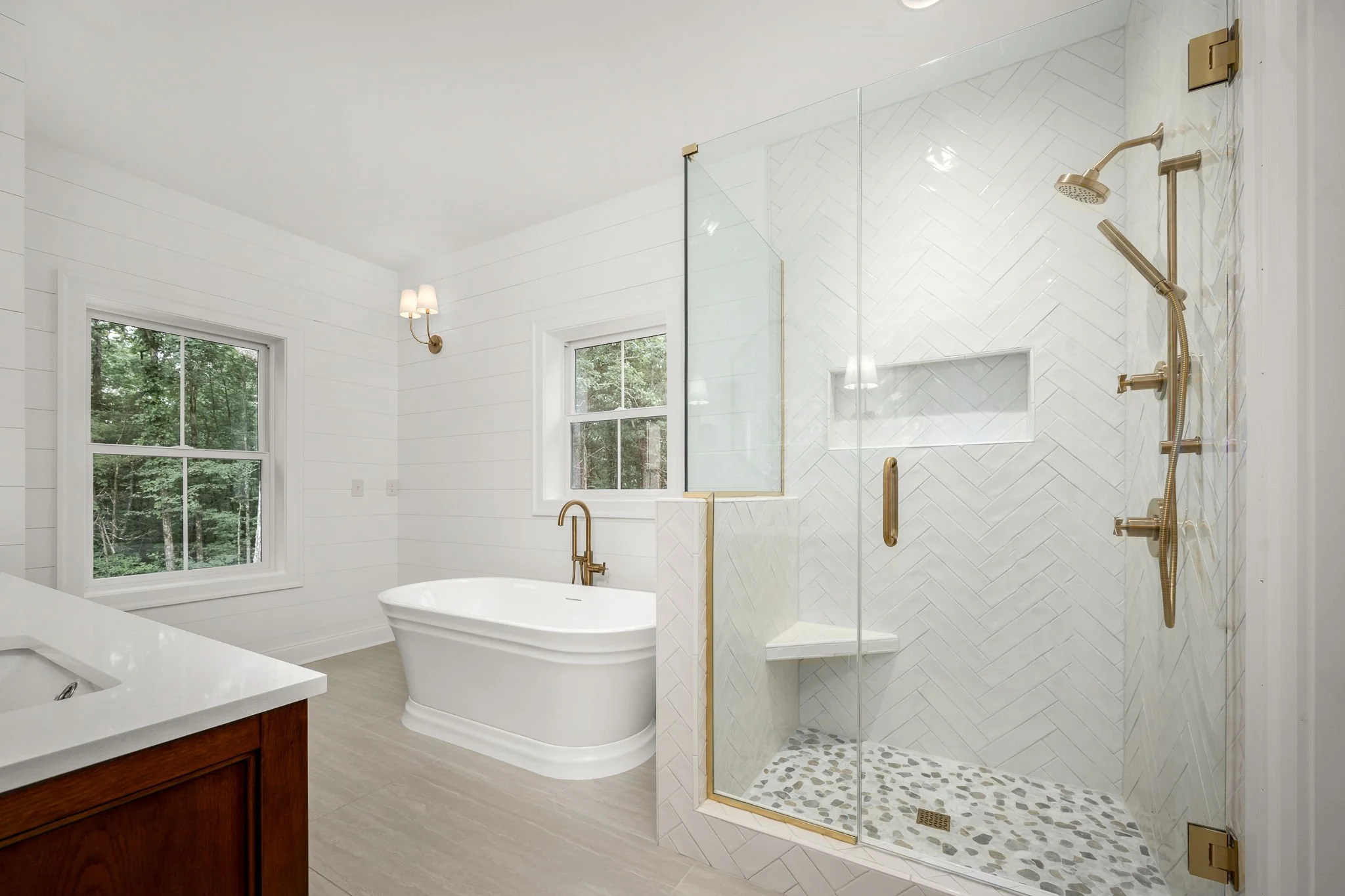 A modern bathroom with a large white bathtub beneath two windows, a walk-in glass shower with gold fixtures, and white shiplap walls.