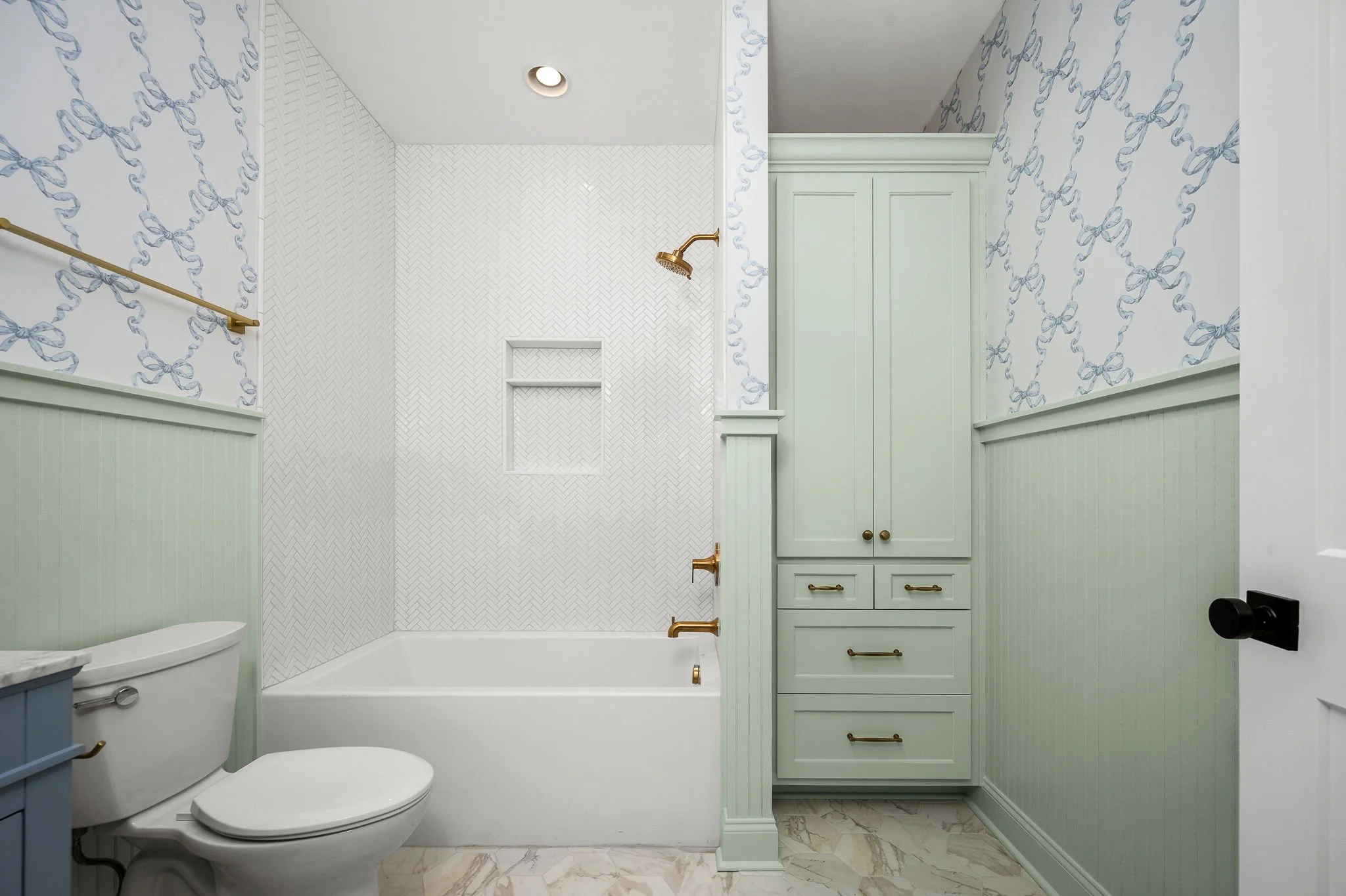 A bathroom with light green wainscoting and cabinets, a white bathtub with brass fixtures, a white toilet, and patterned wallpaper. There is a small round ceiling light and a brass showerhead.