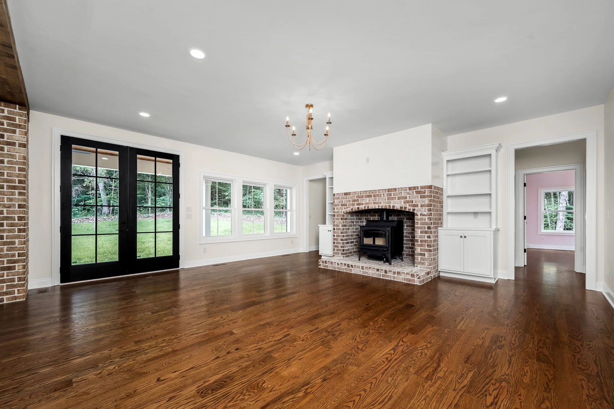 Interior view of a living room with large windows, a brick fireplace with a wood stove, white built-in shelves, hardwood floors, and a brass chandelier on a white ceiling.