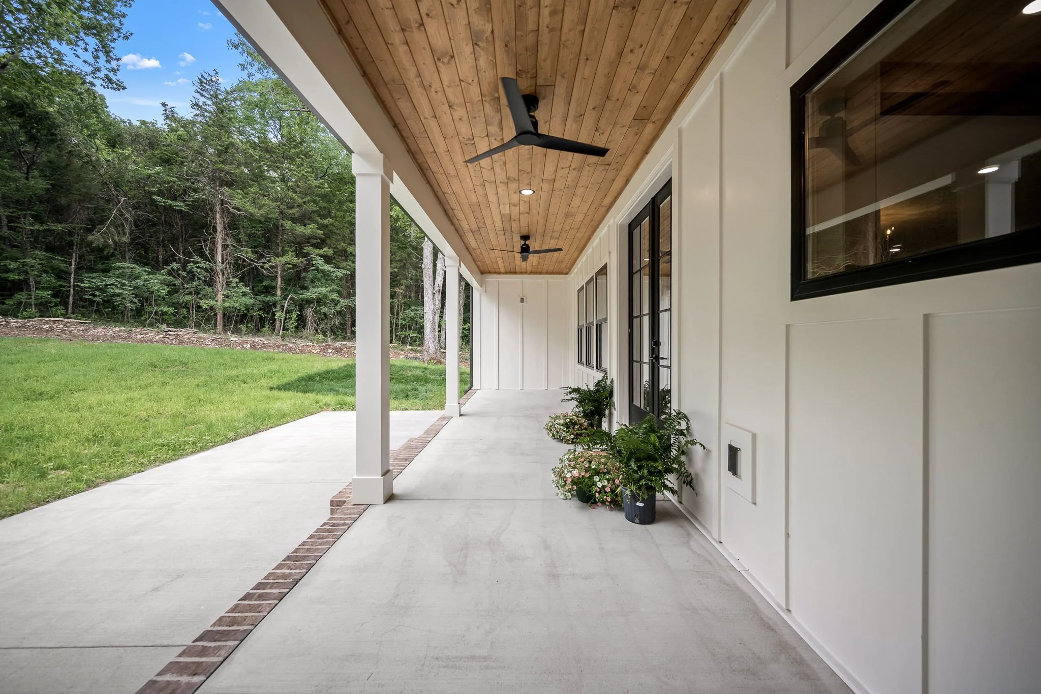 Covered patio with wooden ceiling, ceiling fans, and potted plants outside a house, with a view of a grassy yard and trees.