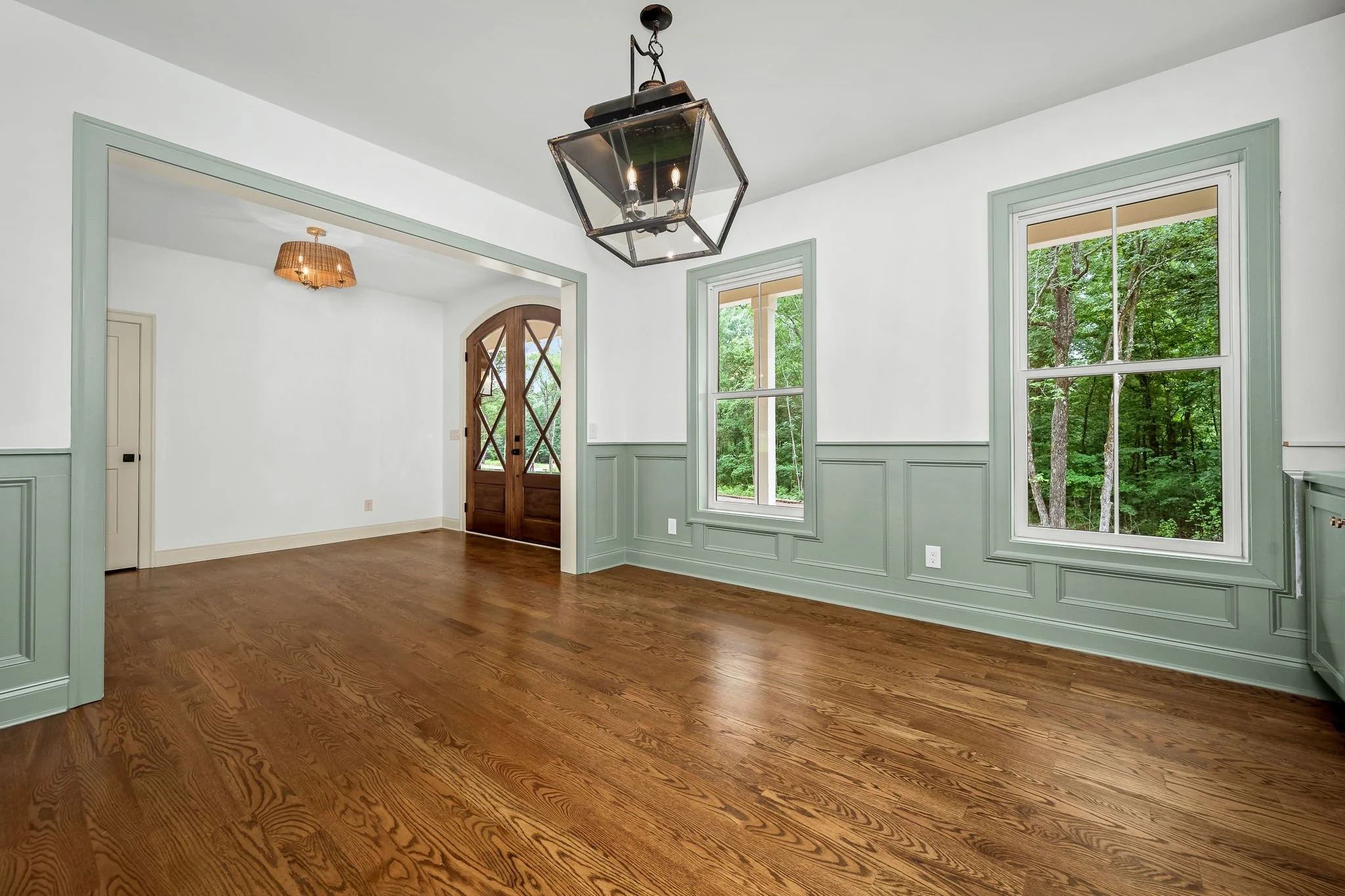 Empty living room with hardwood floors, large windows looking out to green trees, and a wooden front door. Walls are white with light green trim. Ceiling has a black metal light fixture and a smaller wall-mounted lamp.