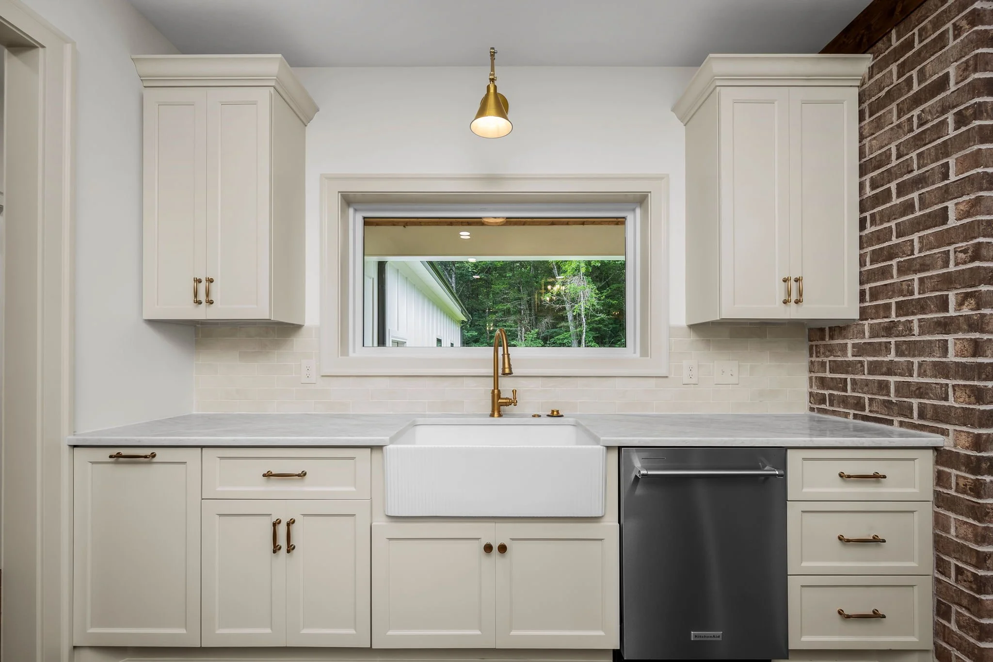 Kitchen sink with a window, white cabinets, brass hardware, a brass faucet, a brick wall on the right, and a white marble countertop.