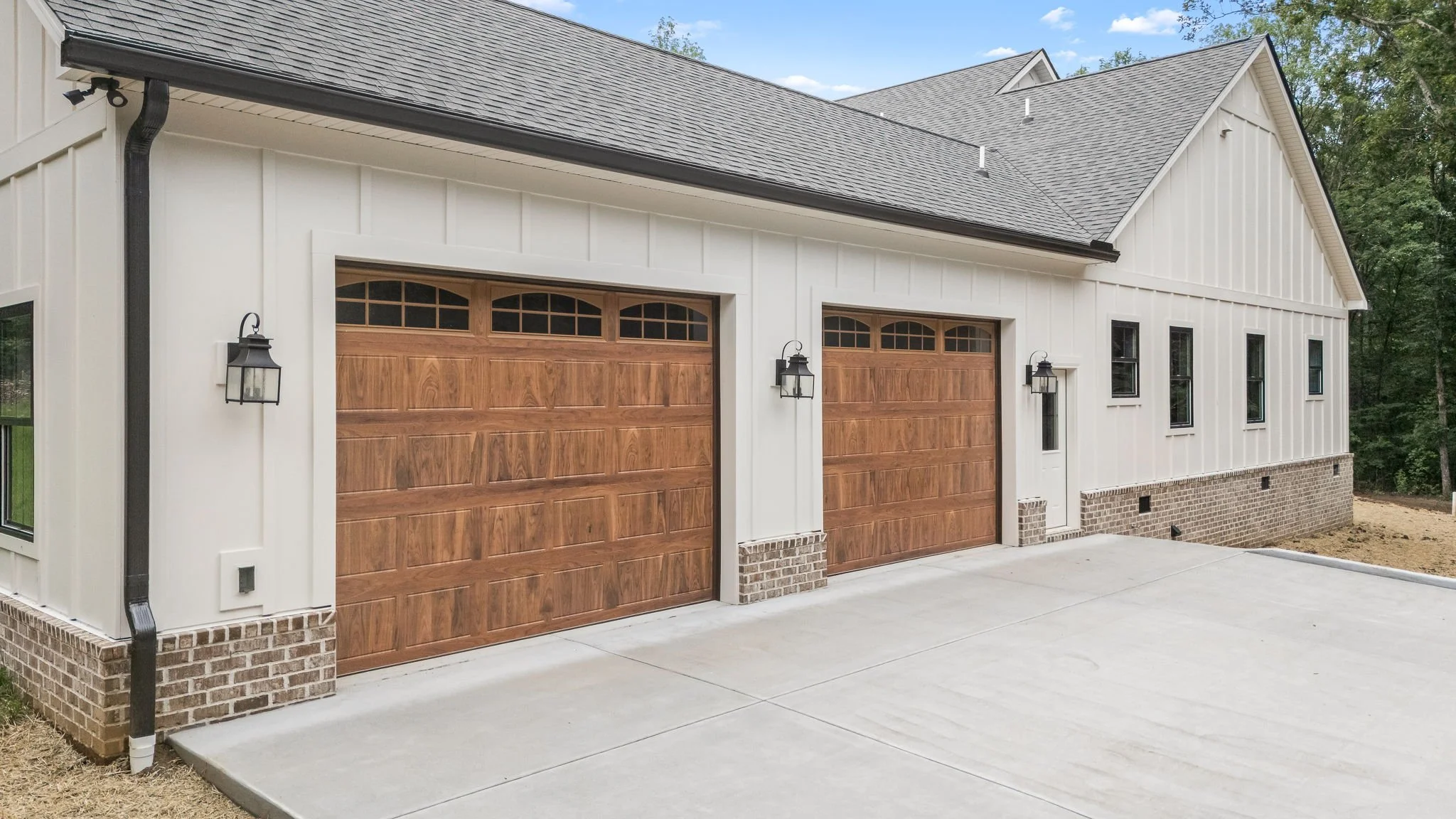 Front exterior of a modern house with two wooden garage doors, black lantern-style wall lights, a brick foundation, and a concrete driveway, surrounded by trees under a blue sky.